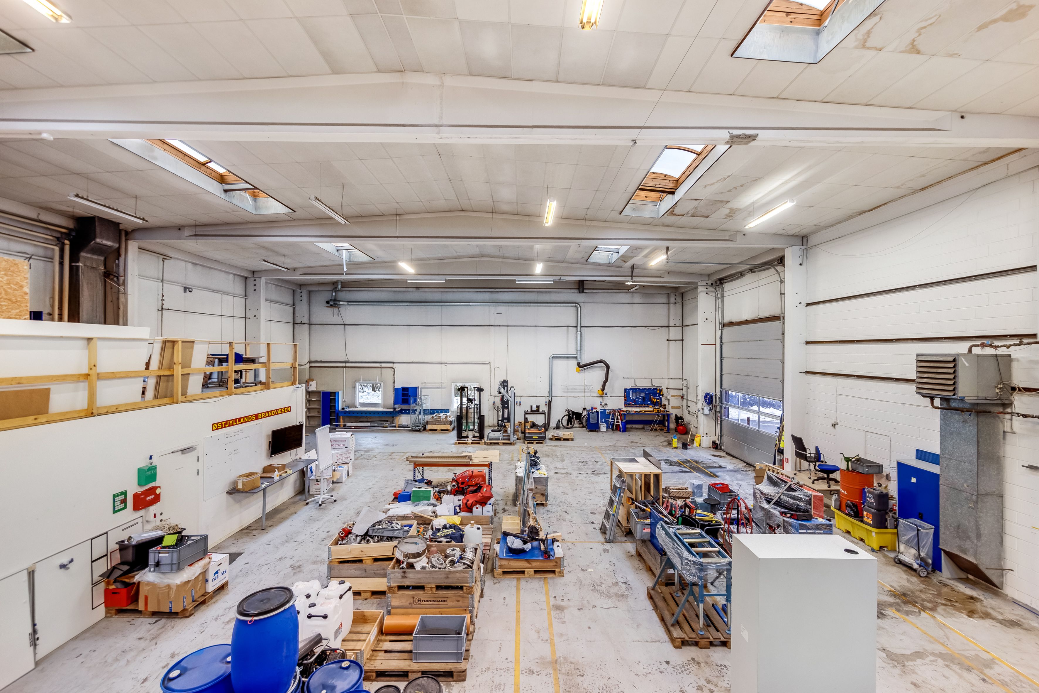 Højloftet lager med lyse skifertage, lysekroner og vinduer i loftet. Væggene er hvide og der er flere lagerreoler med