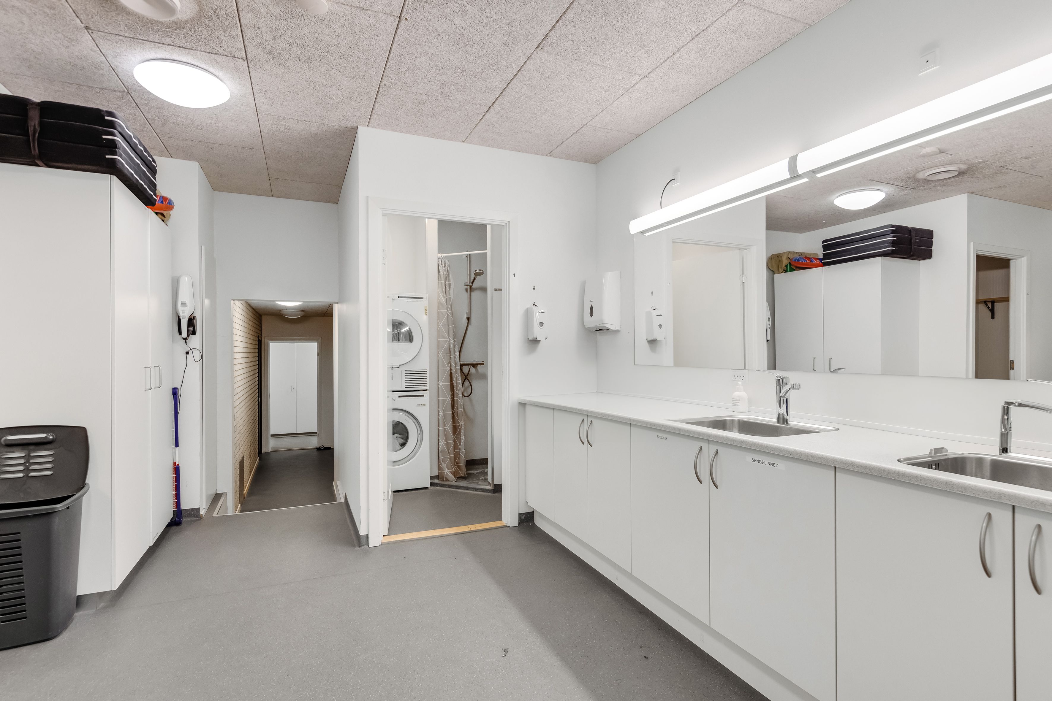 Lager med lyse vægge og ren vaskeri afdeling, med to vaskemaskiner, to spejle og to hængeskabe samt lyse loftslamper