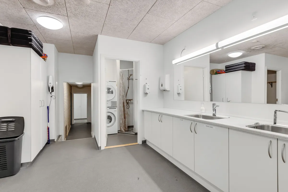 Lager med lyse vægge og ren vaskeri afdeling, med to vaskemaskiner, to spejle og to hængeskabe samt lyse loftslamper