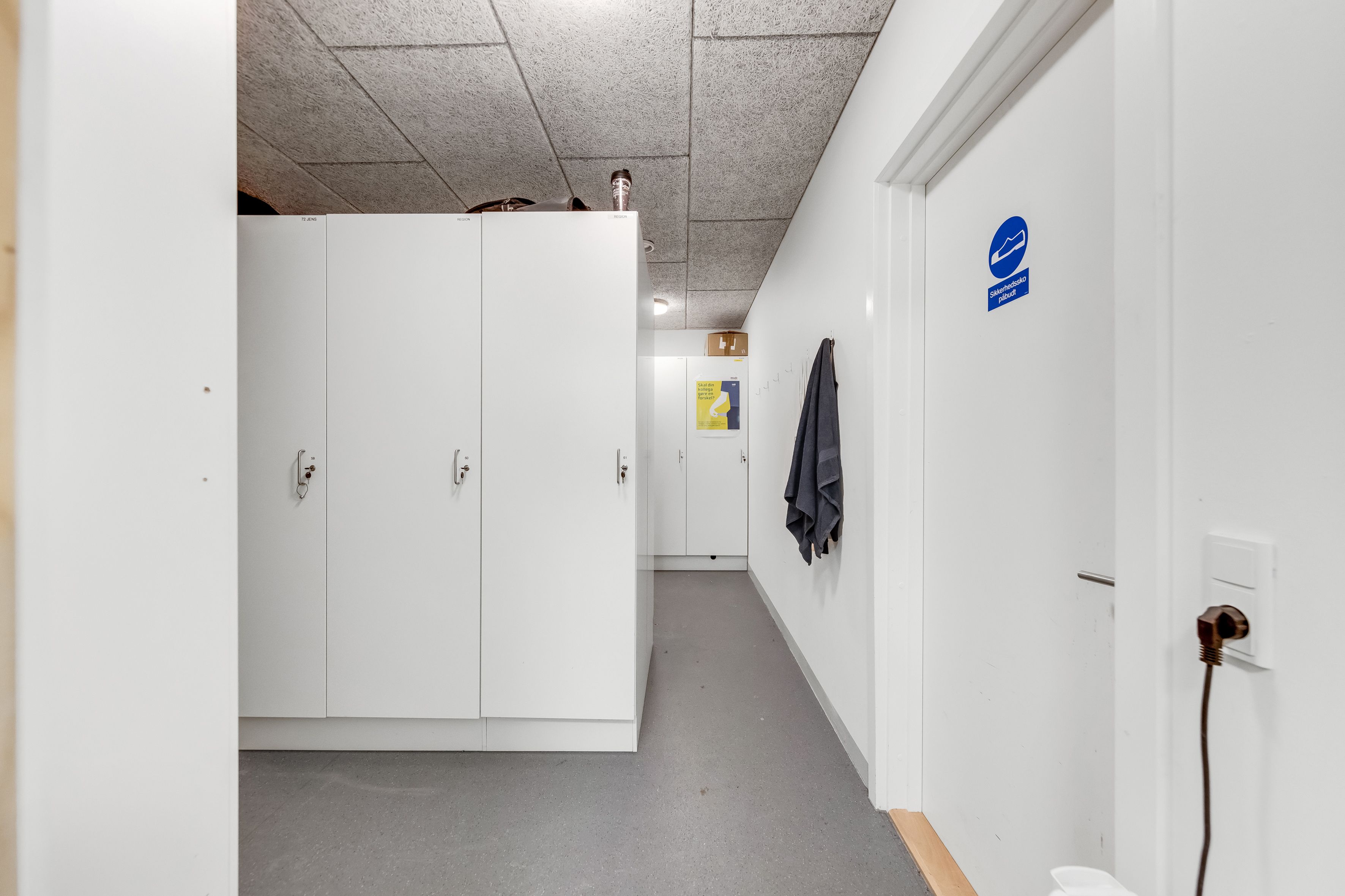 Lager med højloft og lyse loftslampe. Der er hvide kabiner med skilte og en hængende trøje. Loftet er lavet af cement.