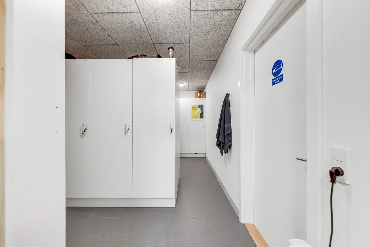 Lager med højloft og lyse loftslampe. Der er hvide kabiner med skilte og en hængende trøje. Loftet er lavet af cement.