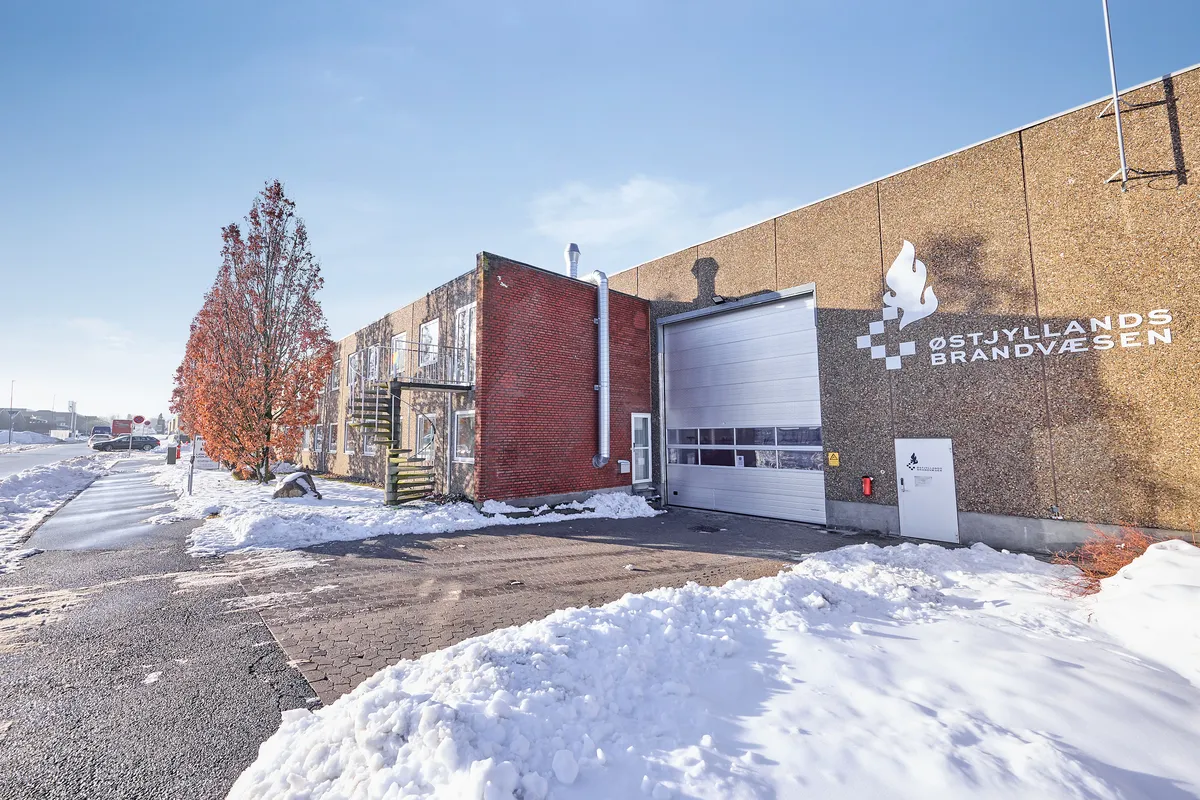 En stor bygning med røde mursten og et hvidt garagedør. Foran bygningen er der en stor snebank og en vej, hvor der er