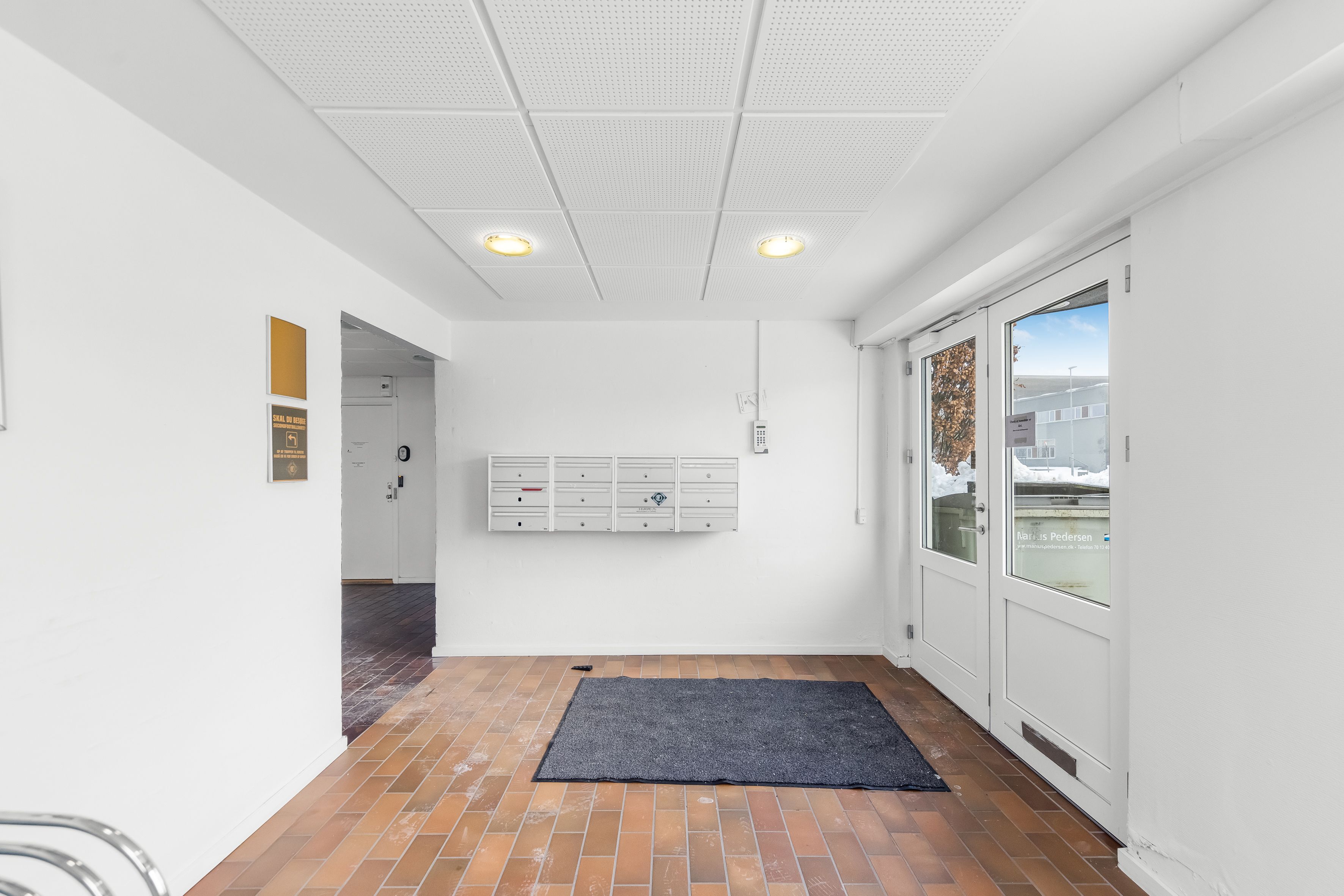 Kort gang med lysegul loftslampe og trægulv. En dør fører til et lagerrum med postkasser og en anden dør til et udendørs