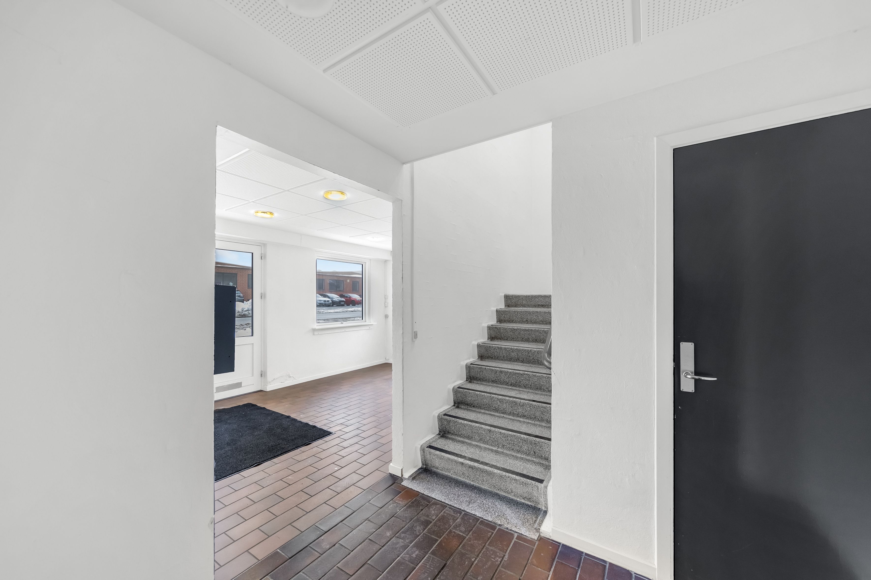 En hvid hall med trægulv og en stigning med hvide og grå trin og en sort dør med sølvfarvet håndtag