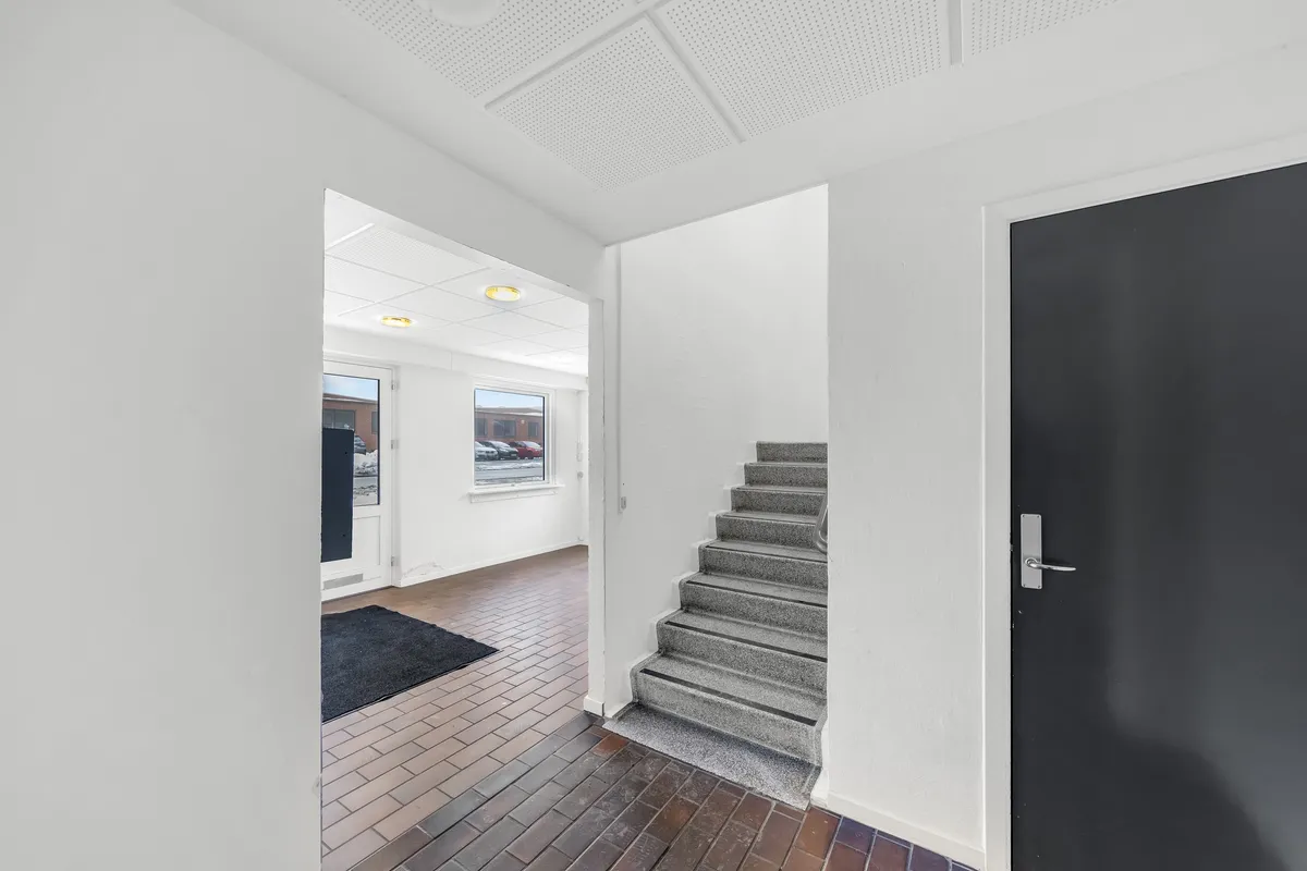 En hvid hall med trægulv og en stigning med hvide og grå trin og en sort dør med sølvfarvet håndtag