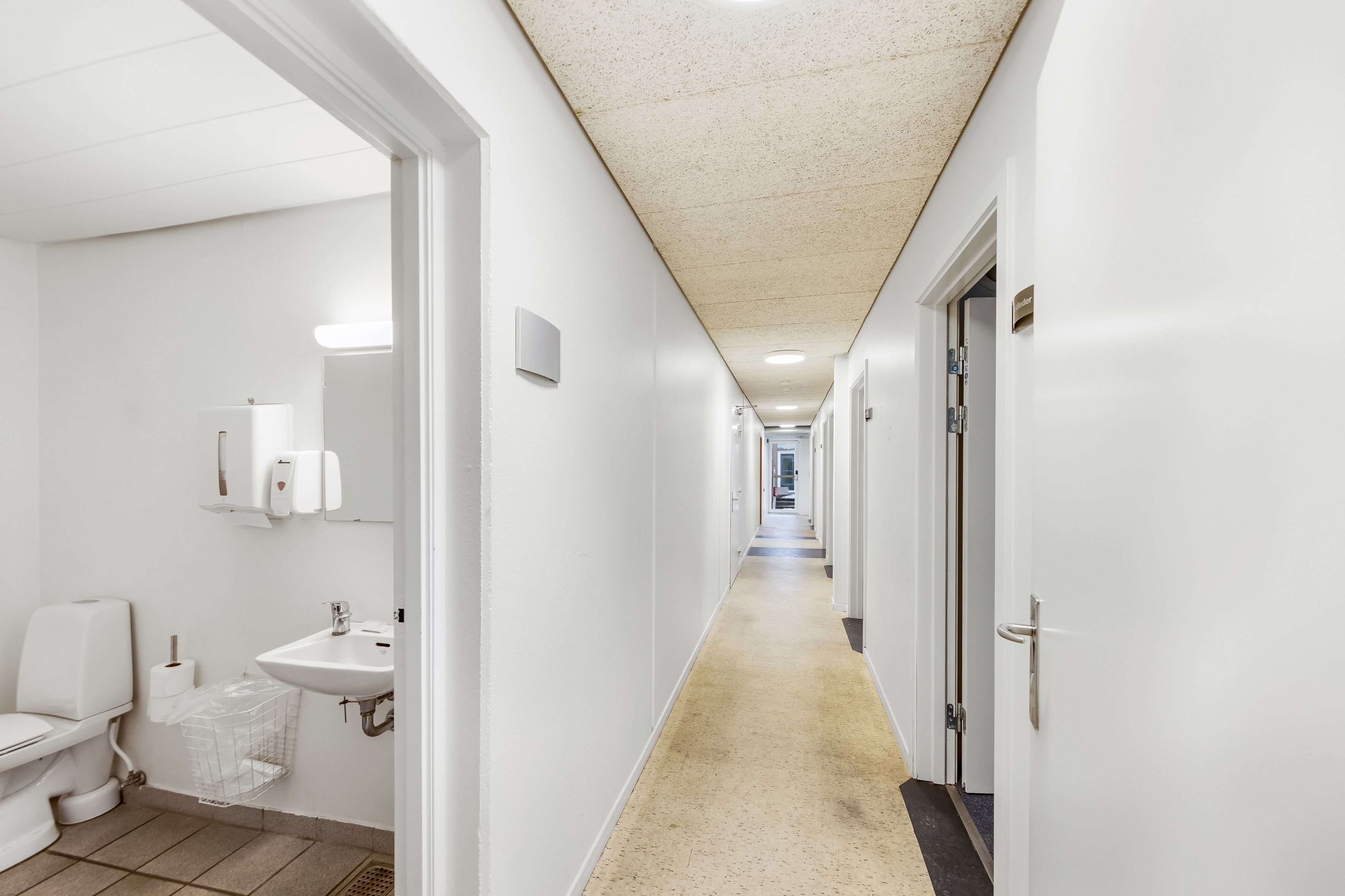 Hvidt og rent toilet med lukket dør og toiletpapir i kurv. Væg og loft er hvide, og der er gulvbelægning i gulbrunt.