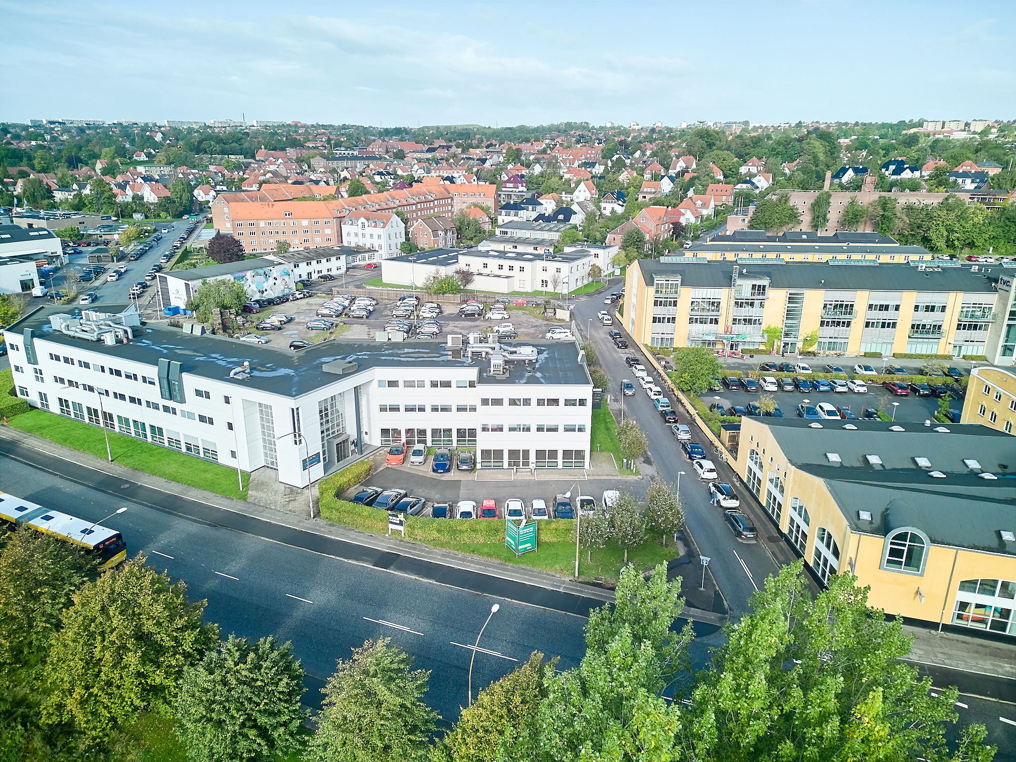 Busstoppested og parkeringsplads med mange biler parkeret. Moderne bygning med hvid facade og flere etager. Få huse og