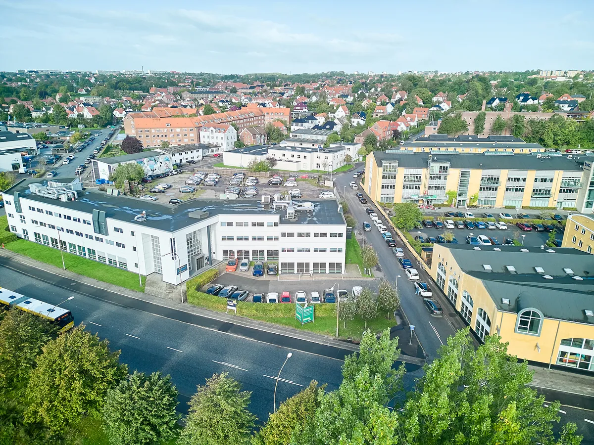 Busstoppested og parkeringsplads med mange biler parkeret. Moderne bygning med hvid facade og flere etager. Få huse og