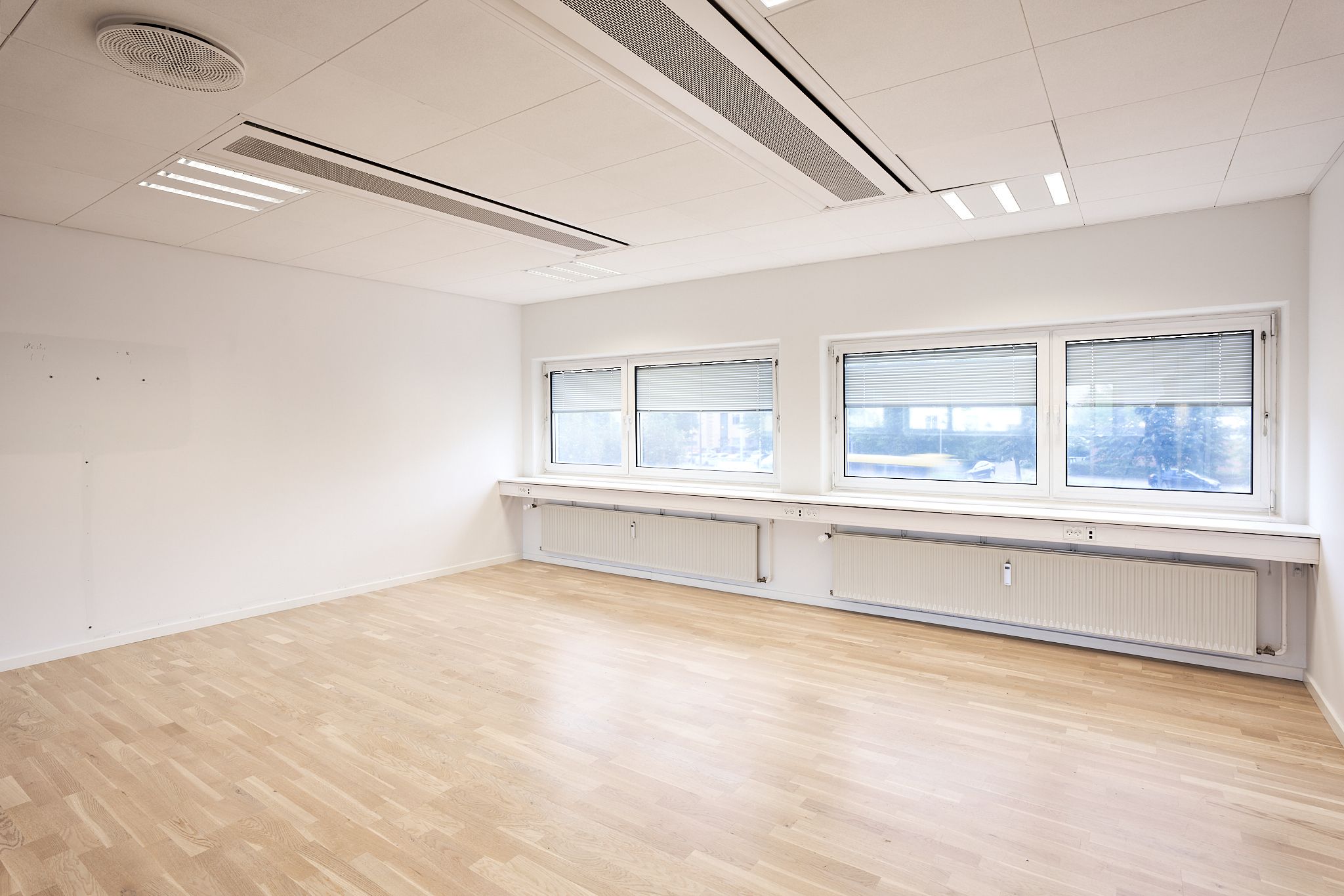 Moderne kontor med hvidmalet væg og trægulv, tre store vinduer med jalusi og radiatorer under hver. Hvide loftspaneler med