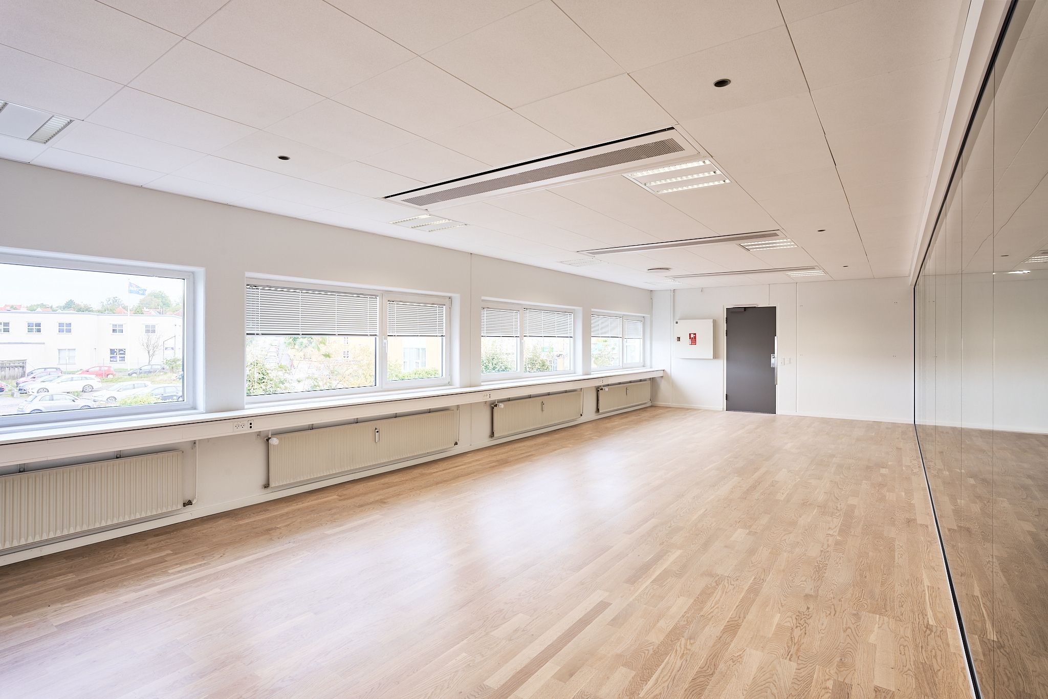 En stor, tom, hvidmalet kontorhal med lyse trægulve, store vinduer med jalousier og integrerede radiatorer langs væggen.