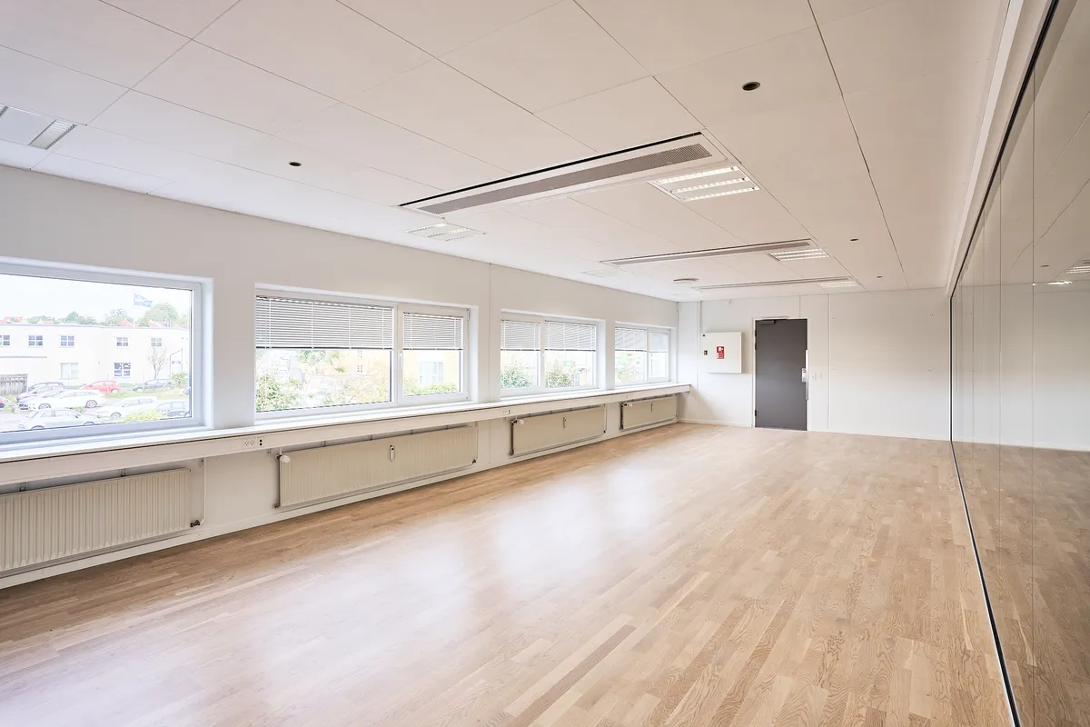 En stor, tom, hvidmalet kontorhal med lyse trægulve, store vinduer med jalousier og integrerede radiatorer langs væggen.