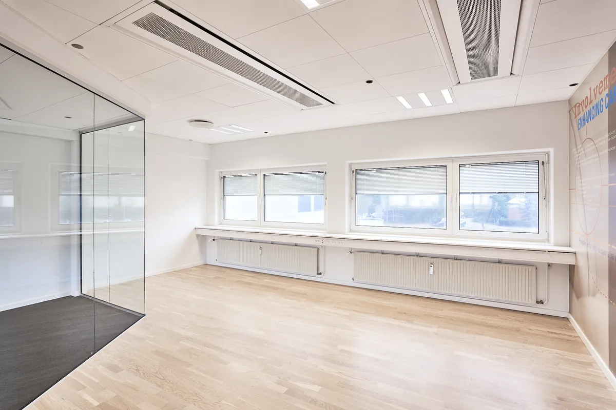Et moderne, tomt kontor med hvide vægge, glasvæg og tre store vinduer med jalousier og undervinduesborde med radiatorer