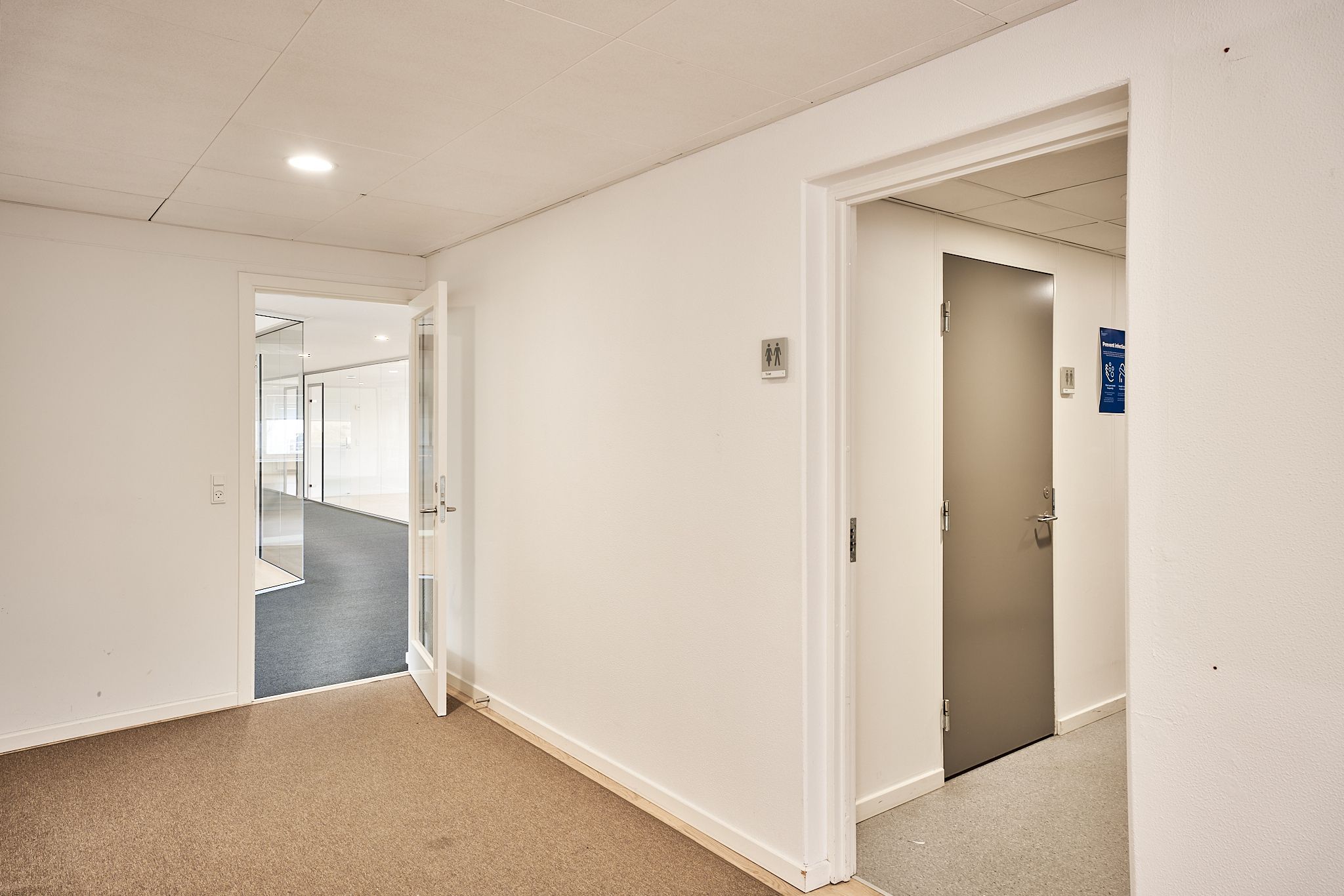 To hvide døre, der fører ind i lokaler med lysegule gulve og hvide vægge med et toiletskilt. En glasdør er åben, og der er