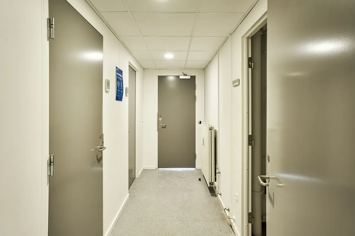 Hallway med hvide vægge, grå døre og en gulvbelægning i gulgrå nuancer. Der er en dør til højre og en til venstre samt en