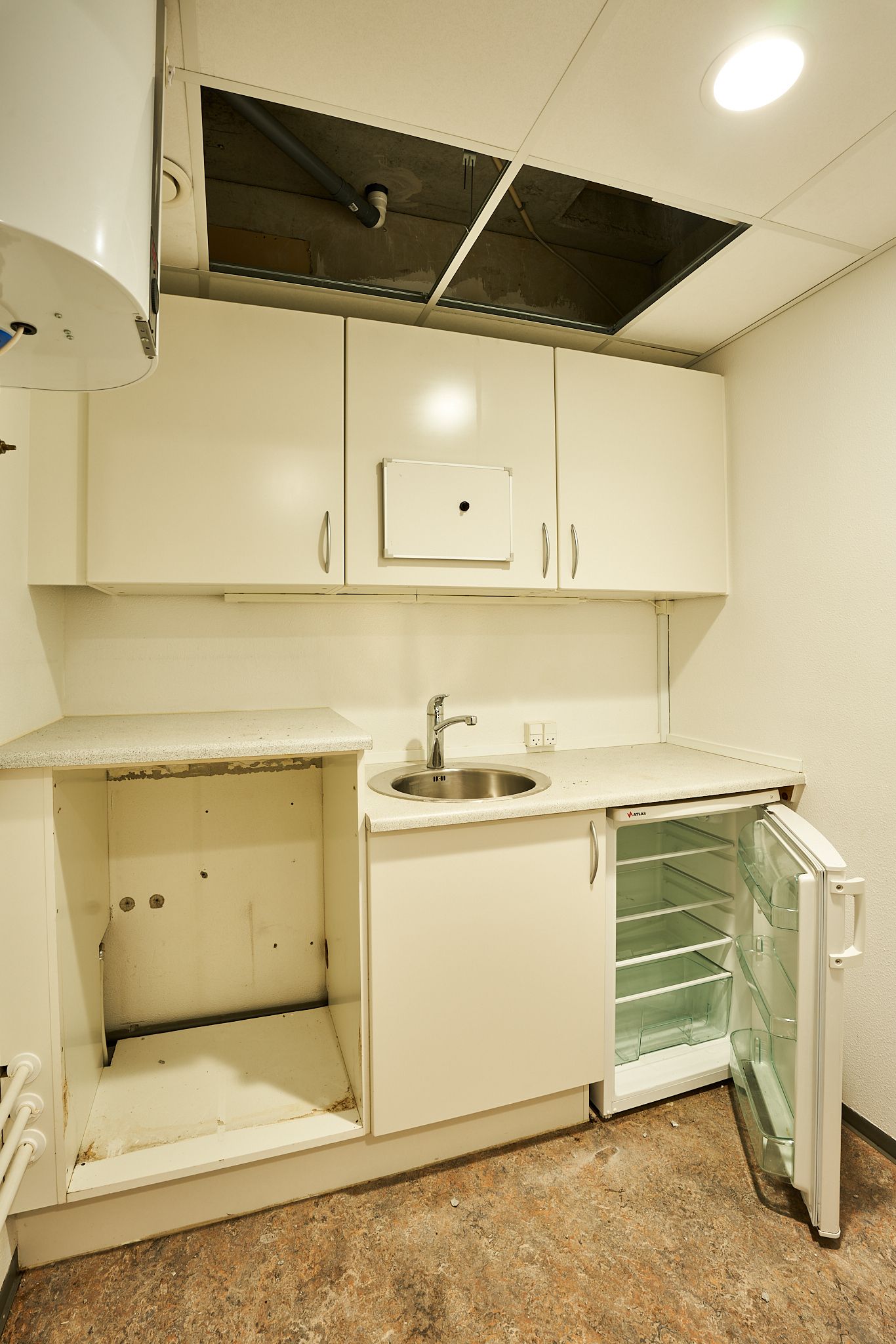 Udenfor køkken er der en hvid køleskab og et rustet vasketøj. Køkkenet er tomt og har et åbent loft, hvor en vandvarmer er