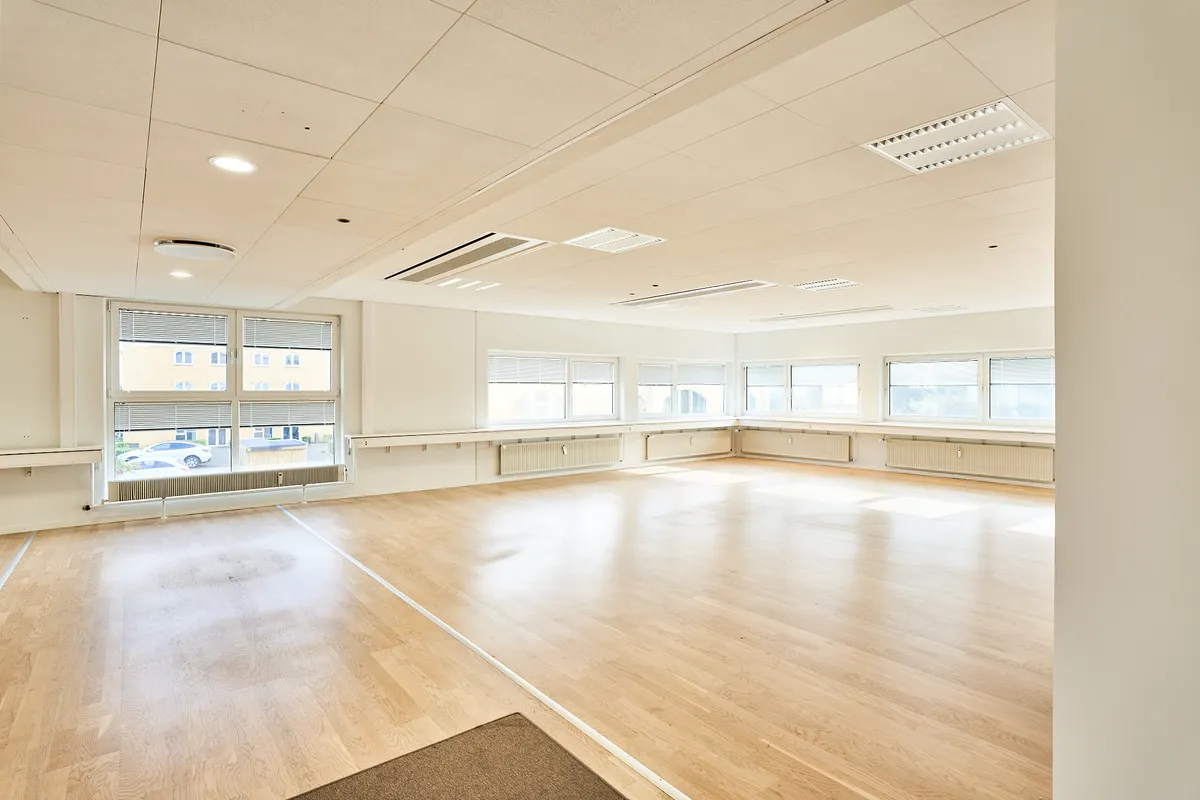 En tom, lys og moderne værelse med hårdt trægulv, hvide vægge og store ruder med jalusierne lukket.