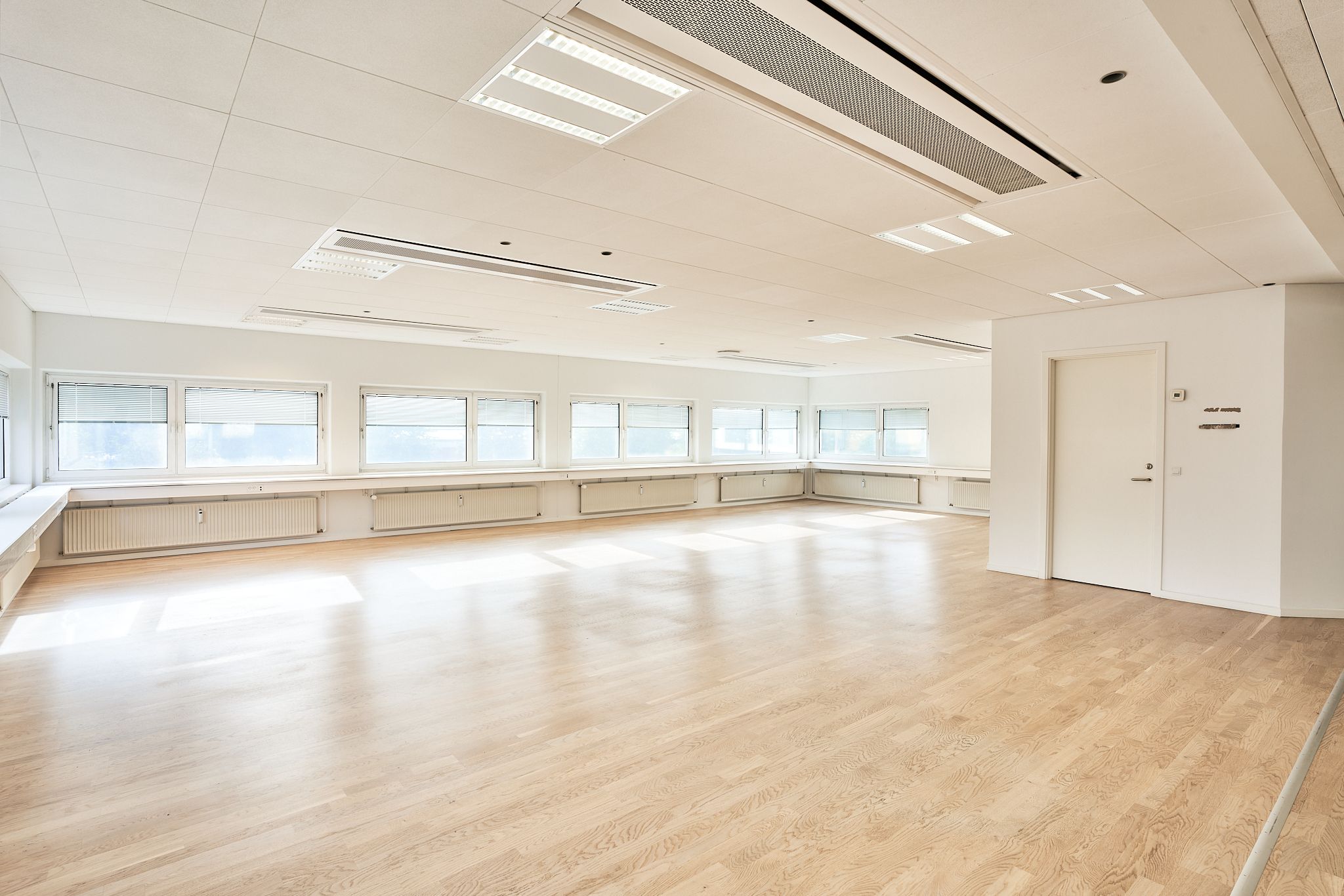 Rummelig lokale med højt loft, hvidmaling, lyse trægulve og store vinduer med gardiner for at give meget naturligt lys