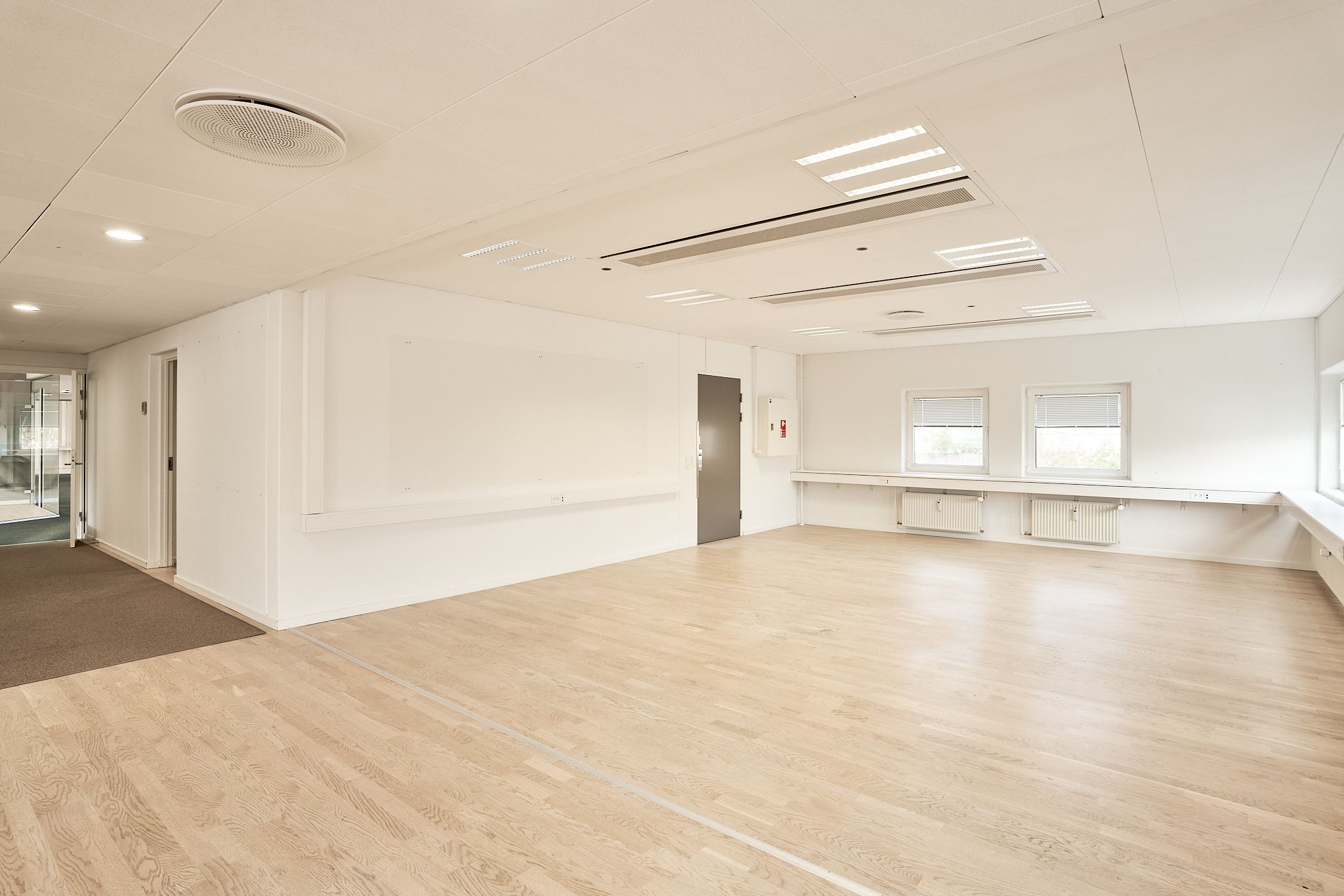 Moderne mødelokale med hvidmalet væg og lyse trægulve. To store vinduer med ruljalusier og radiatorer langs væggen. En