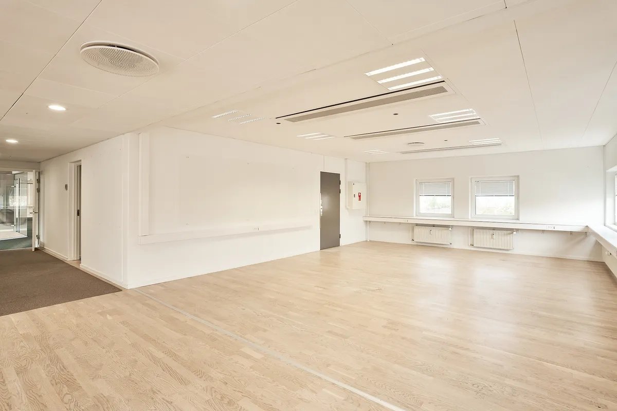 Moderne mødelokale med hvidmalet væg og lyse trægulve. To store vinduer med ruljalusier og radiatorer langs væggen. En