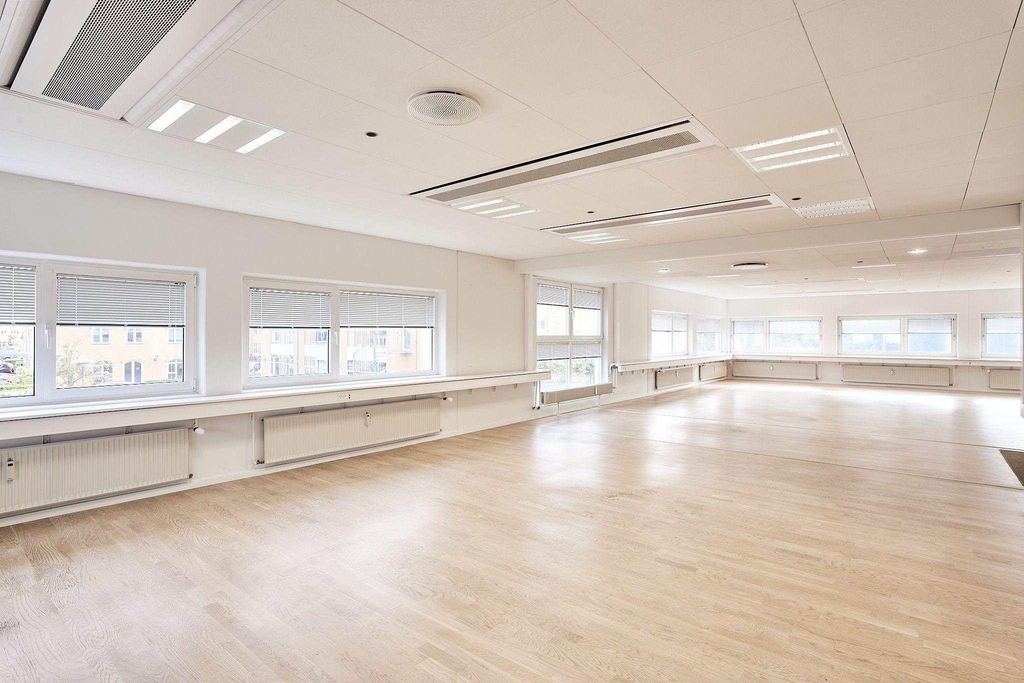 Et større rum med hvide vægge, trægulv og store vinduer med hvide jalousier. Luftventilationer er installeret i loftet.