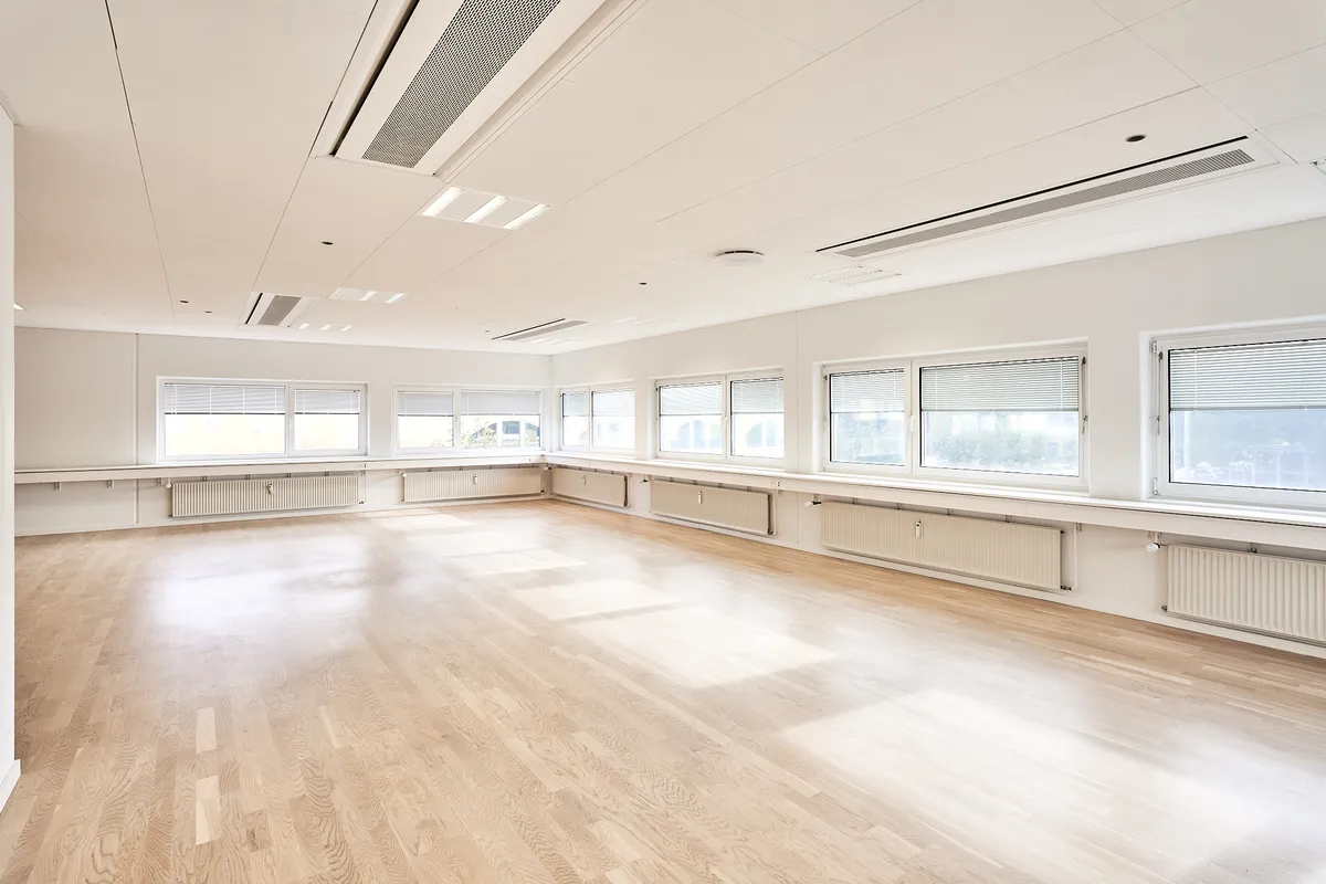 En stor møde- og konferencelokale med hvide vægge, hårdt trægulv, hvide lofter med indbygget klimaanlæg og store vinduer med