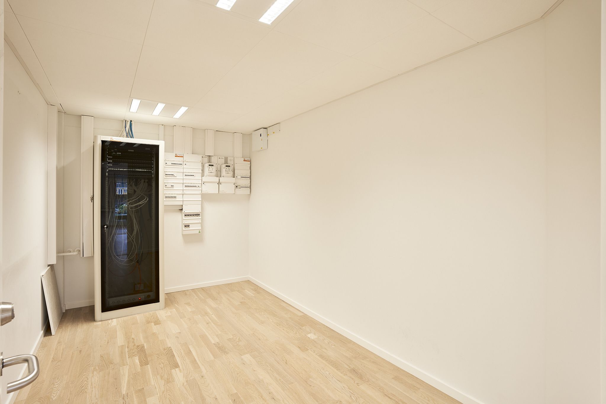 Et lille rum med hvide vægge, højloft og hårdt trægulv samt flere hvide bokse og en stor kabinet til servere og elektronisk