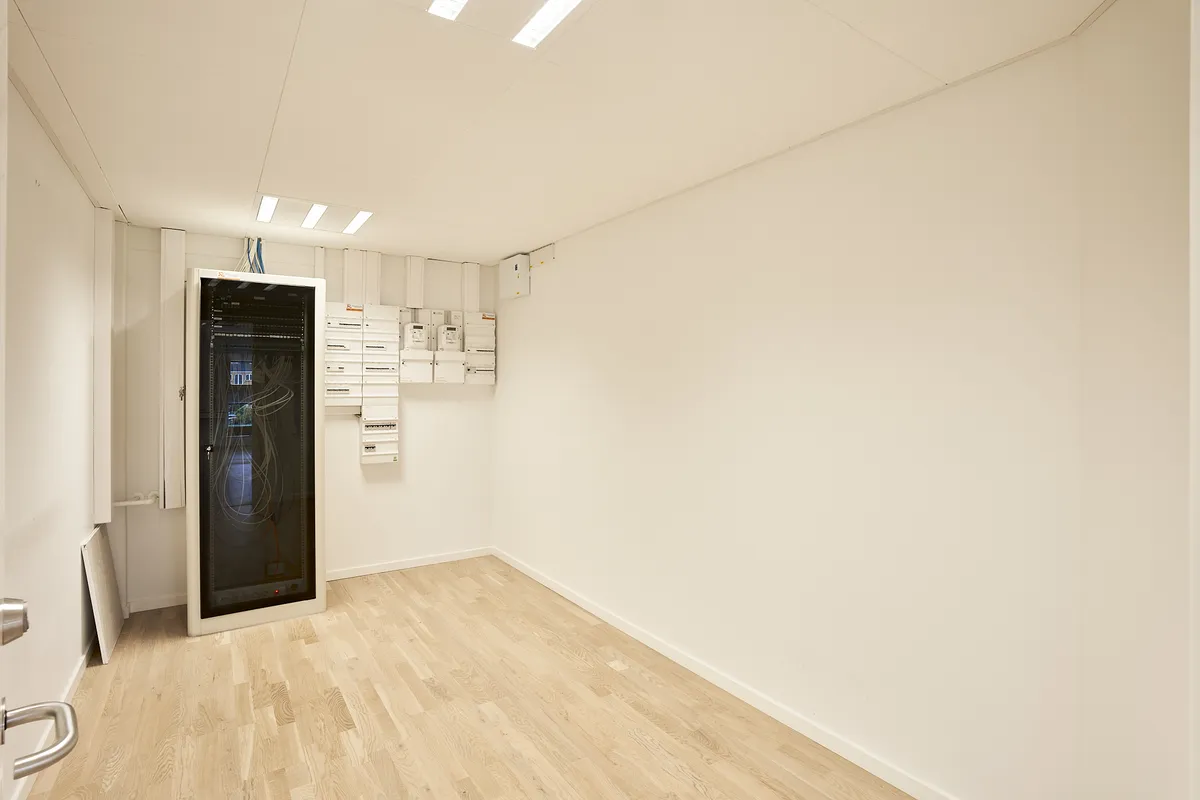 Et lille rum med hvide vægge, højloft og hårdt trægulv samt flere hvide bokse og en stor kabinet til servere og elektronisk