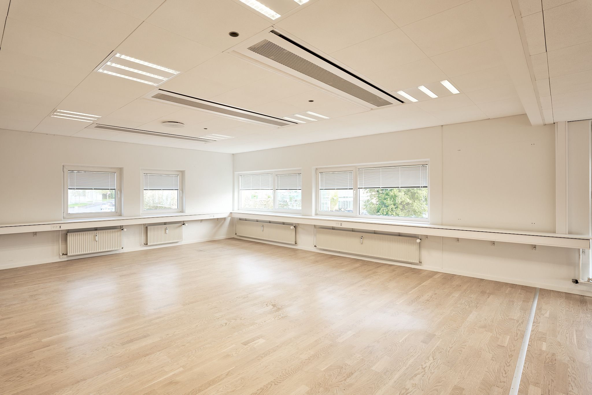 En tom rum med hvidmalede vægge og lyse trægulve samt fire vinduer med lysegardiner, der giver naturligt lys til rummet