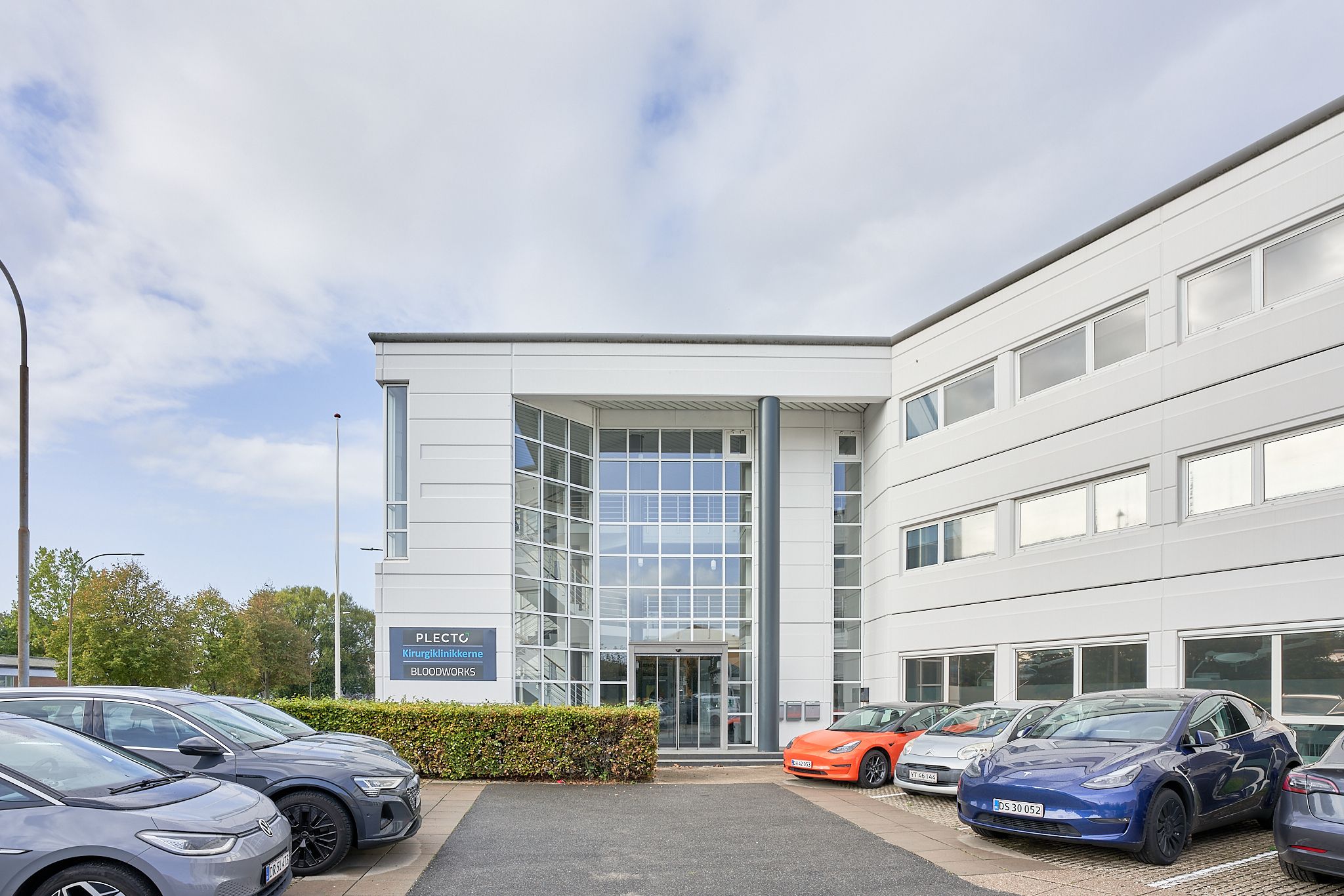 Flere biler parkeret foran et hvidt bygningskompleks med stort glasvinduesfront og skilte med teksten Plectro