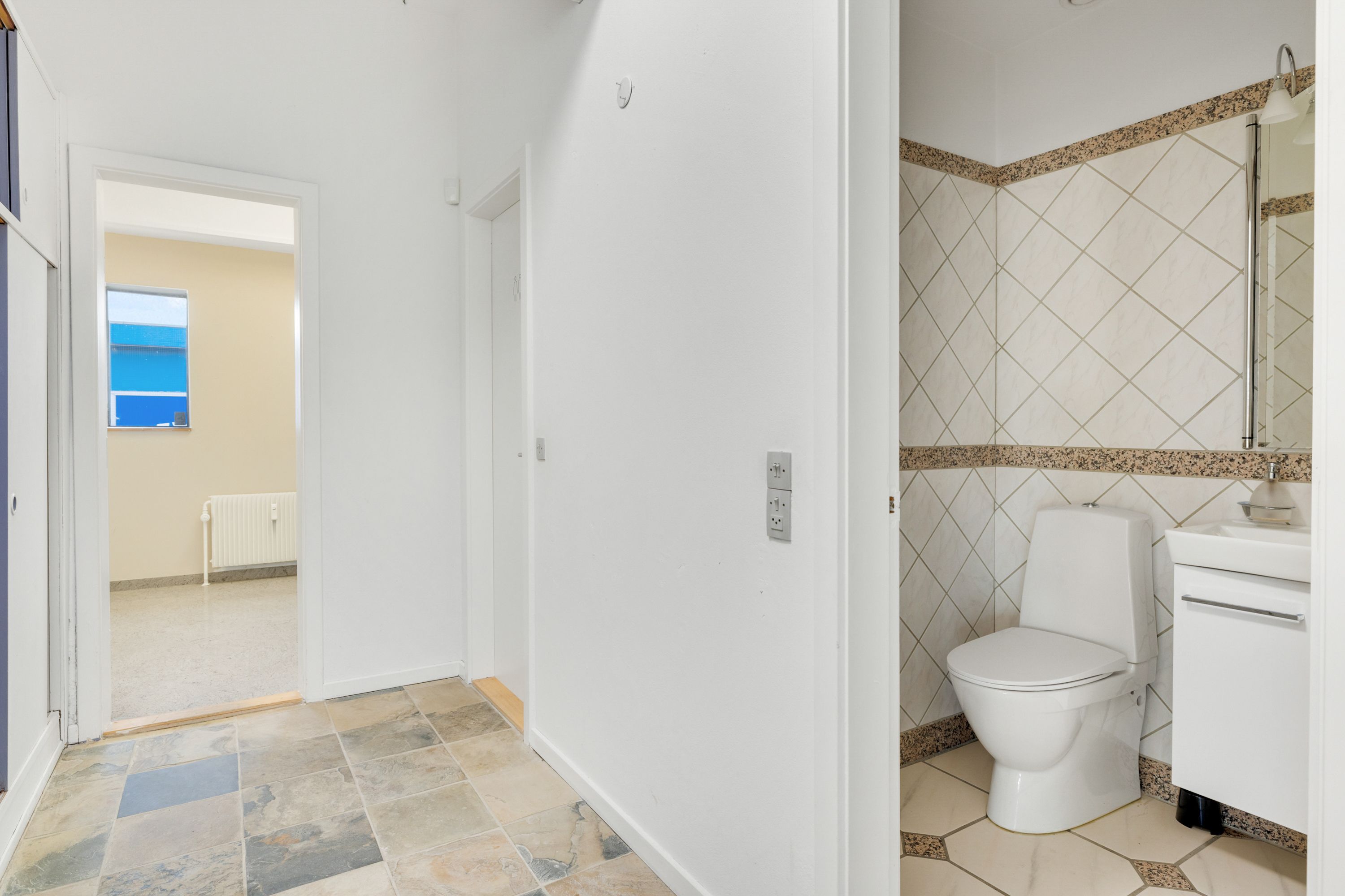 En moderne toiletrum med ren, hvid mur og gulv samt et toilet med hvidt sæde og håndklædeholder, hvor der er et spidset