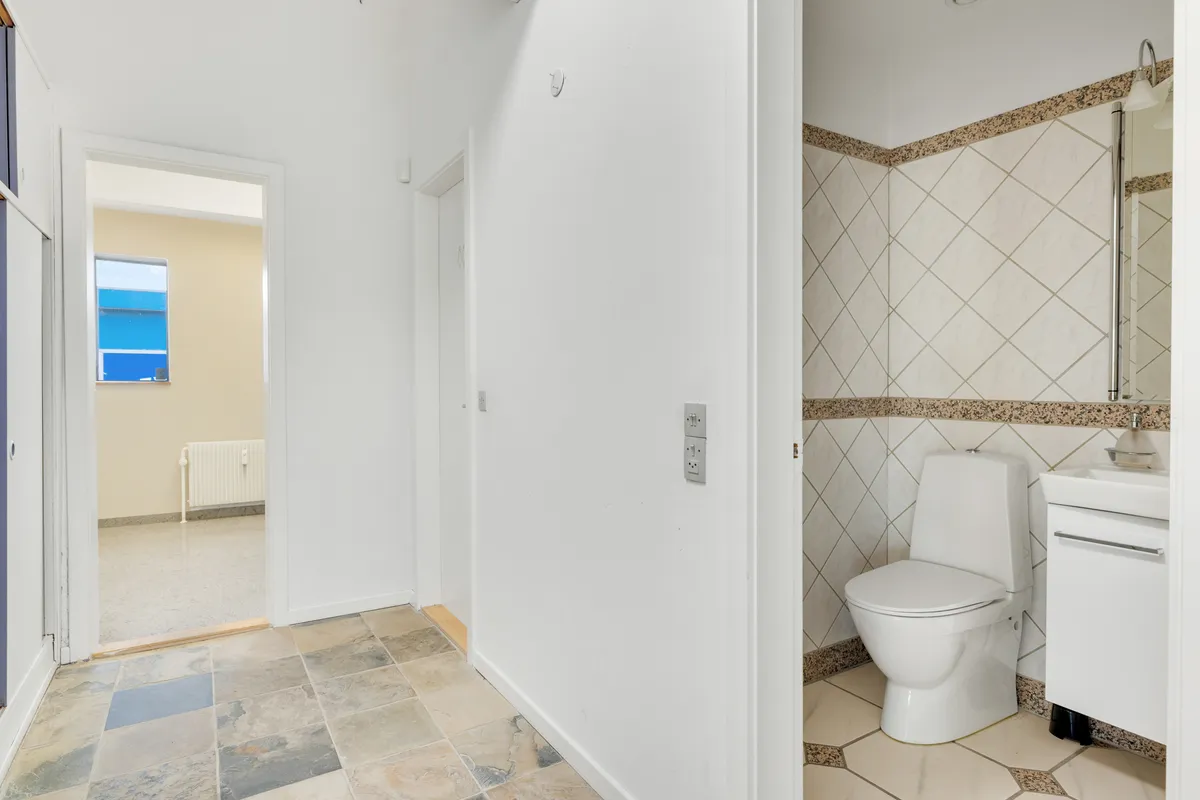 En moderne toiletrum med ren, hvid mur og gulv samt et toilet med hvidt sæde og håndklædeholder, hvor der er et spidset