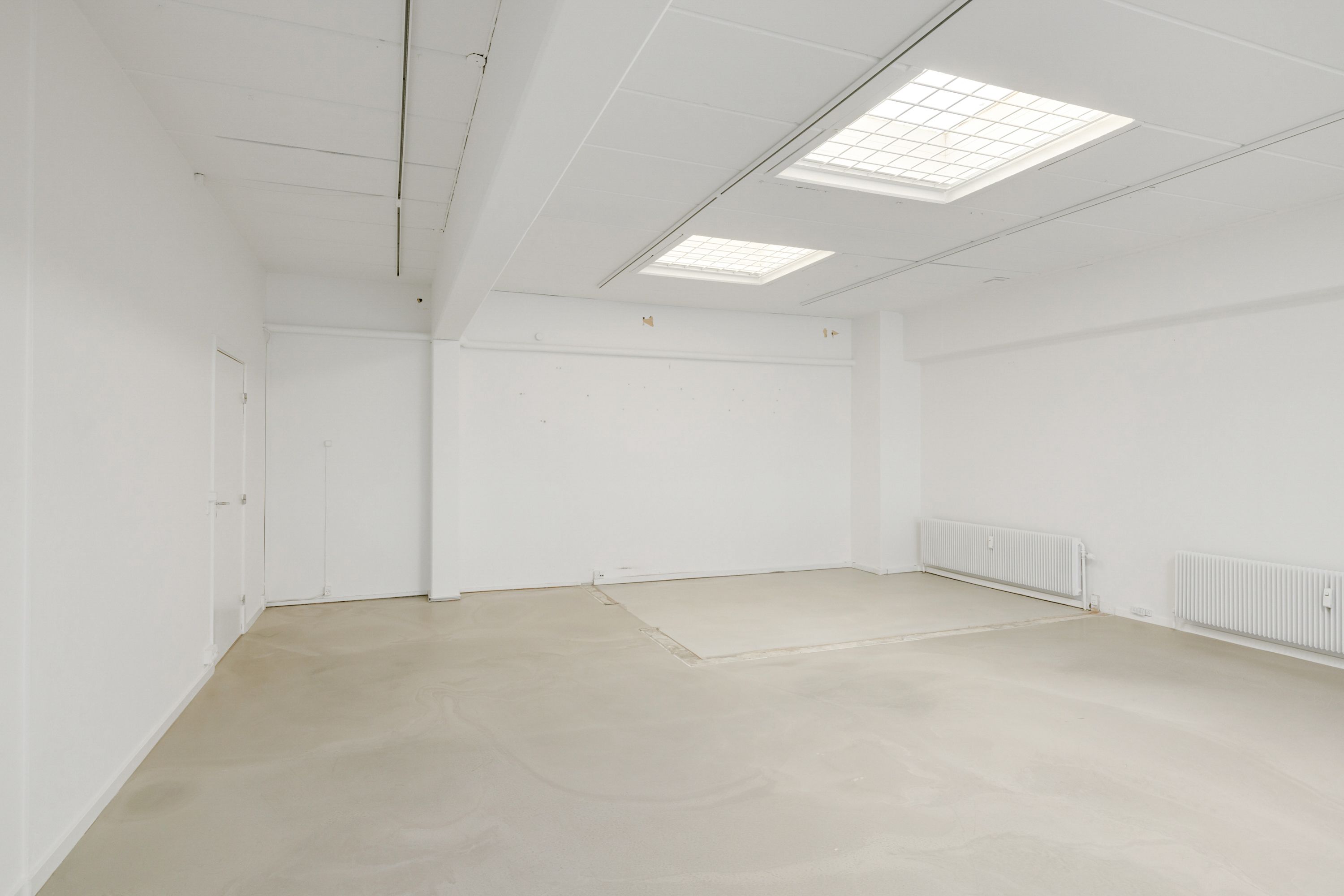 Lokaler med hvidt loft og vægge samt to radiatorer til højre samt to vinduer til loftet og to døre på den ene side