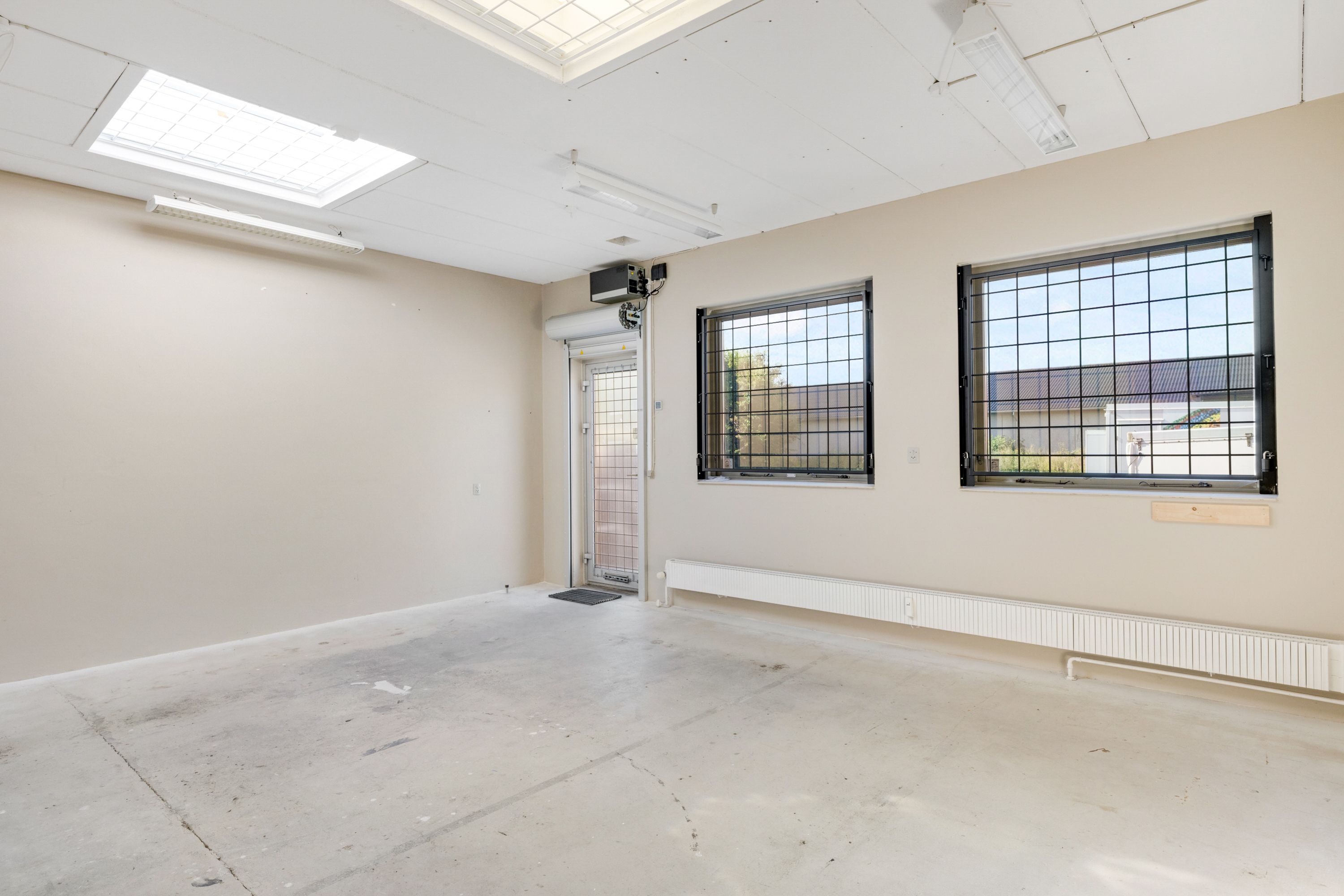 Et tomt lokale med lyse gulve, hvide vægge og to store ruder med metalliske rammer, samt et glasdør med metallisk dørhåndtag