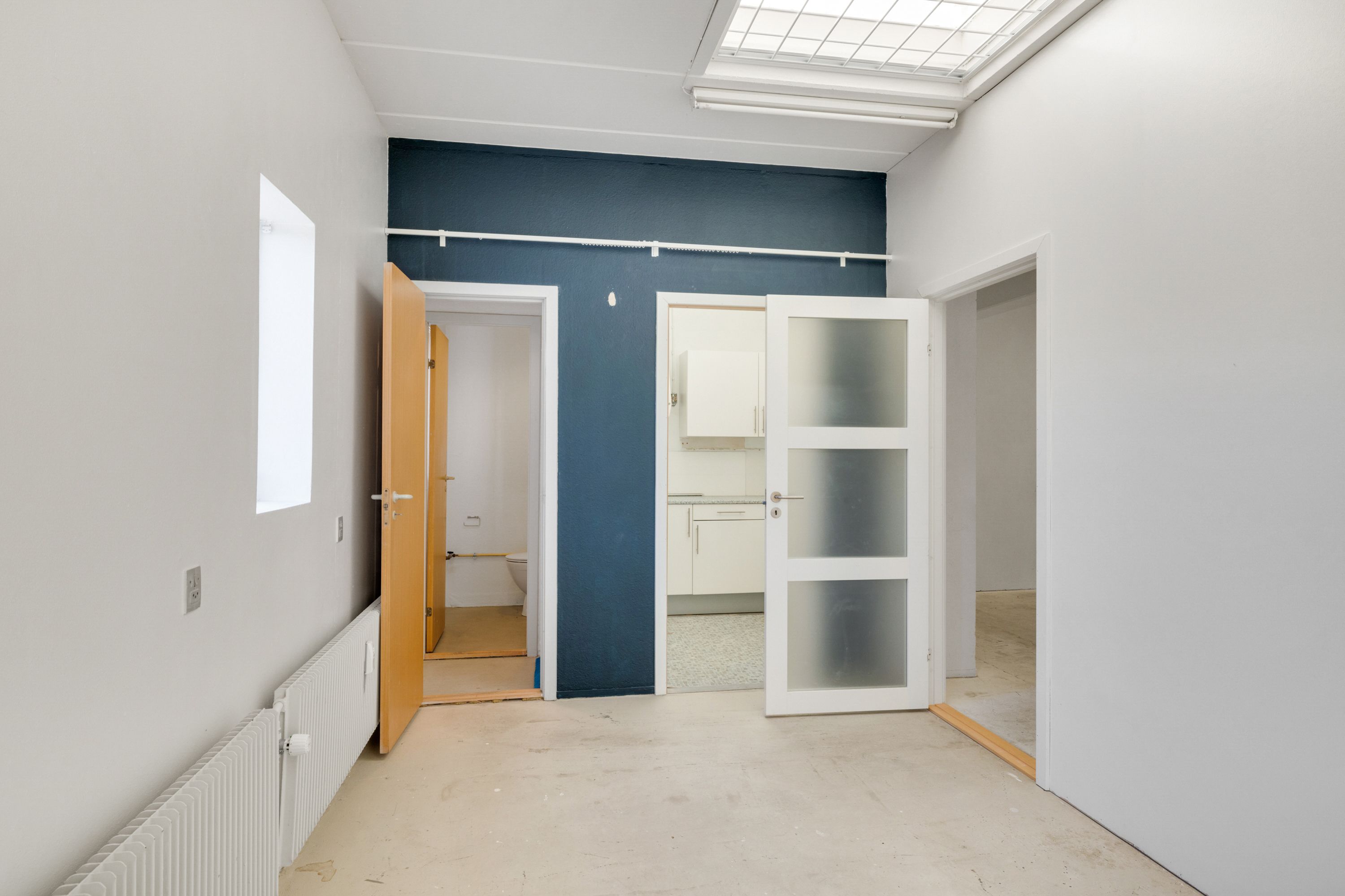 Et moderne kontor med åbne døre og lyse loftsvinduer. En glasdør og en trædør med håndtag. En hvid radiator og en toilet i