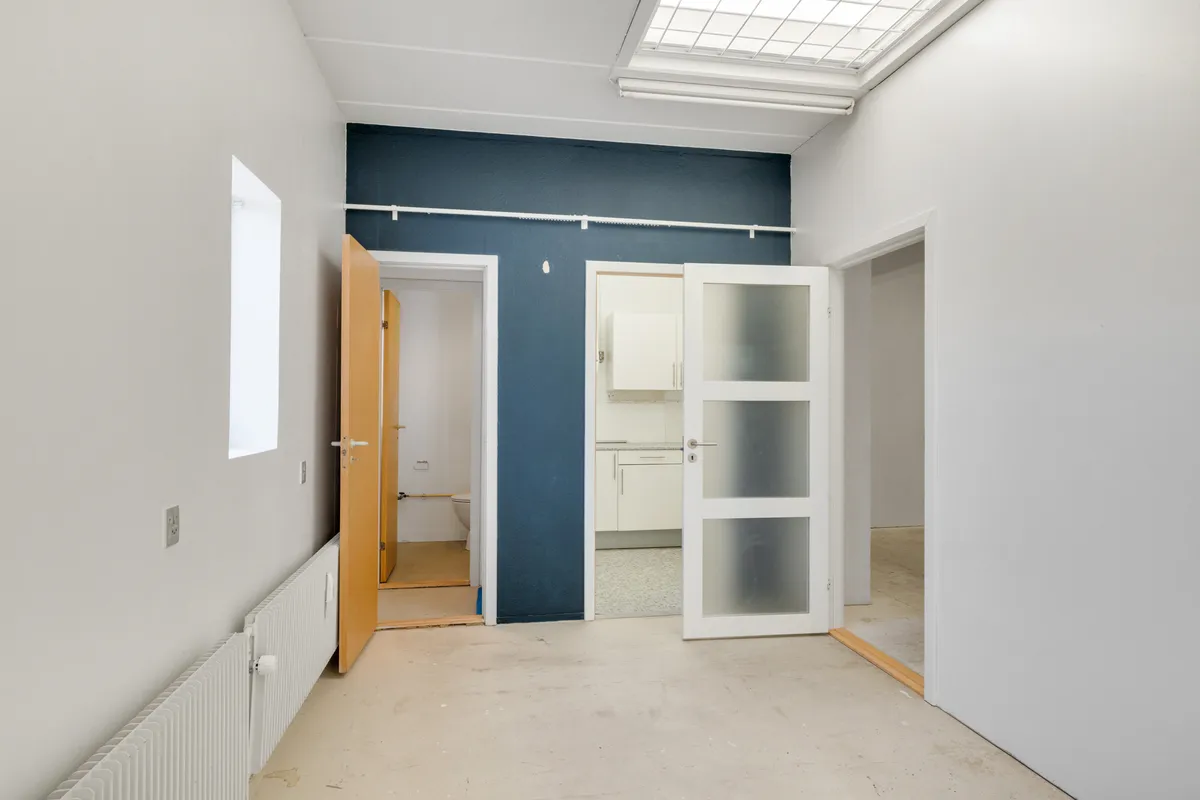 Et moderne kontor med åbne døre og lyse loftsvinduer. En glasdør og en trædør med håndtag. En hvid radiator og en toilet i