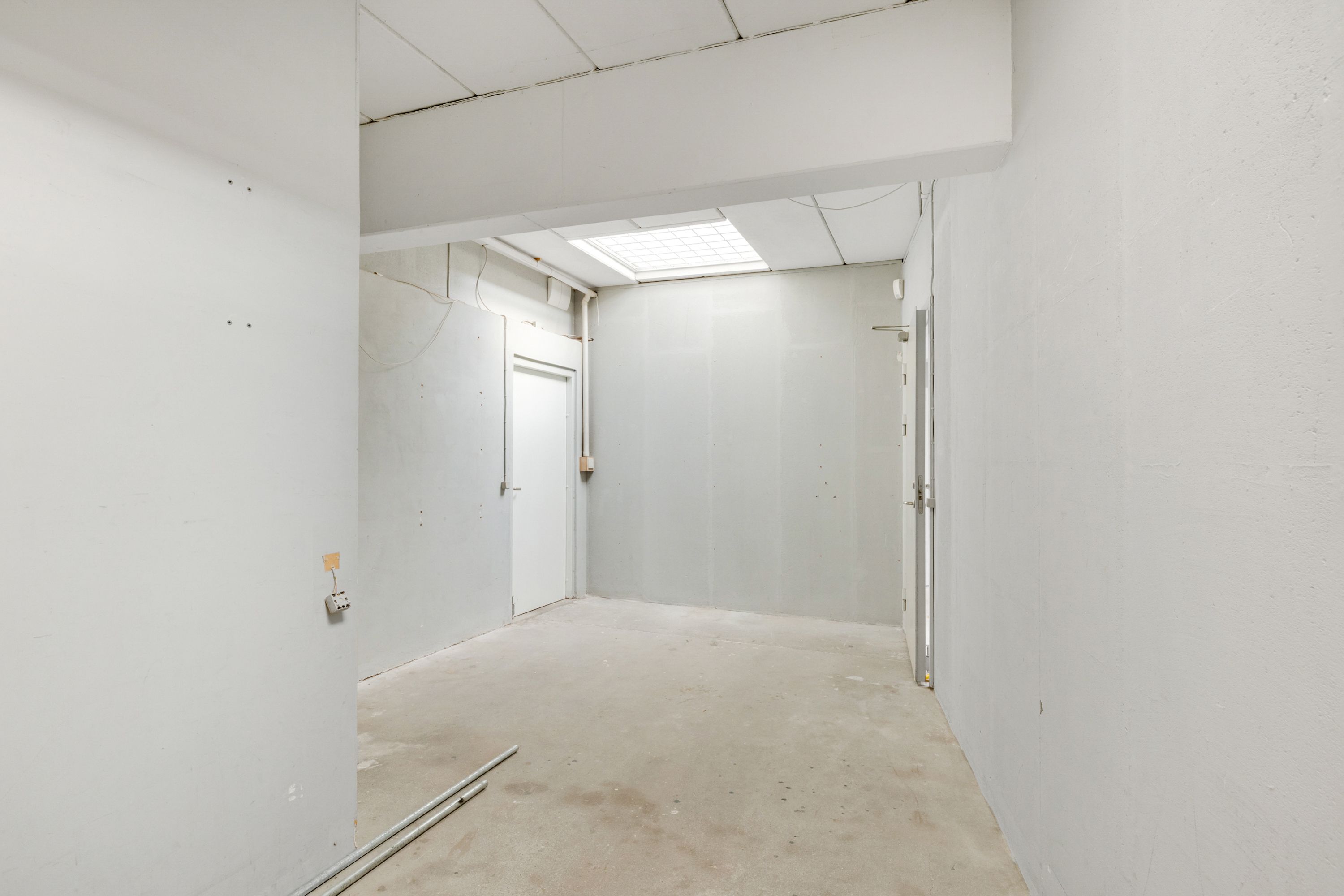 Kort og lyst korridor med hvide vægge og cementgulv, to døre og en dør med beskyttelsesbøjle, og et loft med slanke hvide