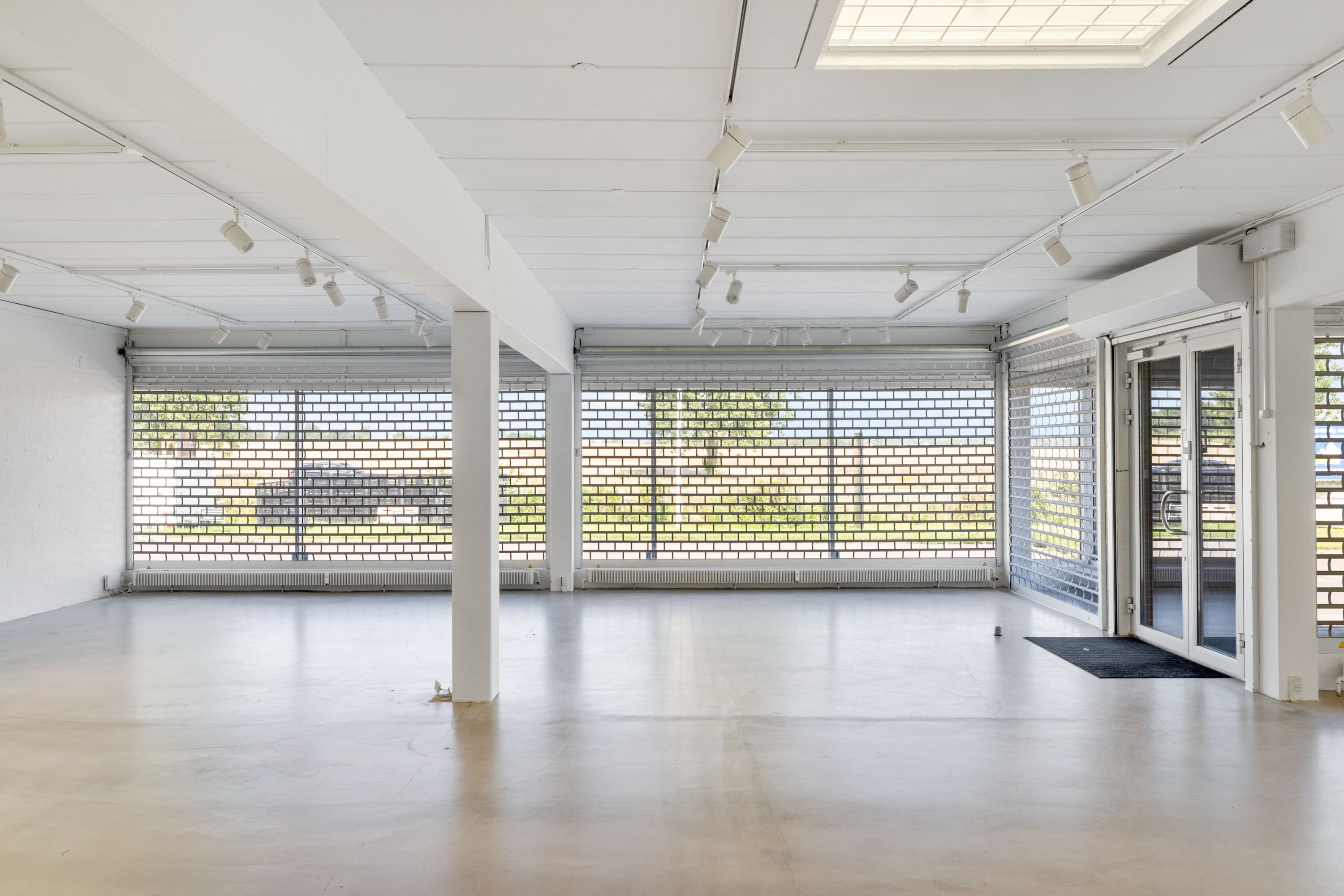 Store lokaler med hvide vægge og gulv, flere vinduer med glasruder, flere spotlys og en hvid dør til kontor.