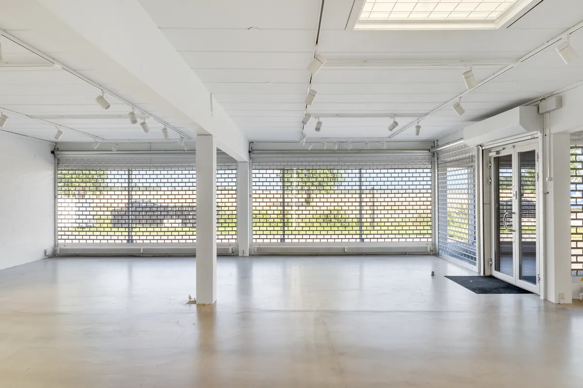 Store lokaler med hvide vægge og gulv, flere vinduer med glasruder, flere spotlys og en hvid dør til kontor.