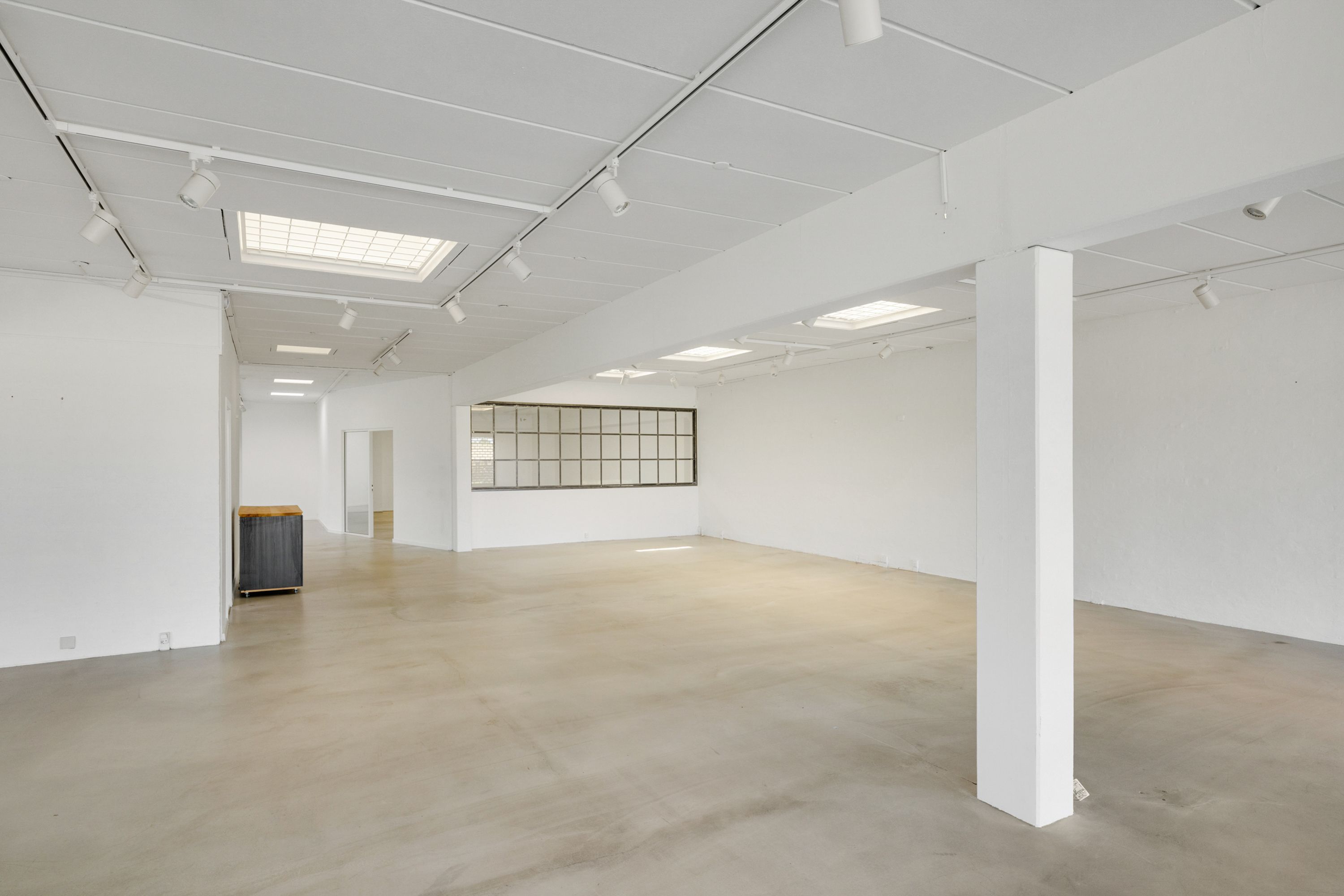 Et tomt og rummeligt rum med hvide vægge og loft samt en hvid søjle og lysekroner, der er ophængt i loftet. Der er en