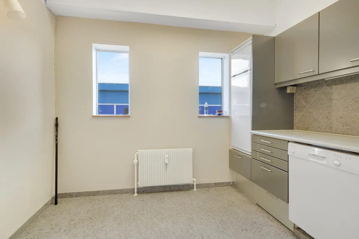 Moderne køkken med lyse vinduerne, hvidt køleskab, lysegrå skabe og ren hvid maskine, der står ved siden af en hvid radiator