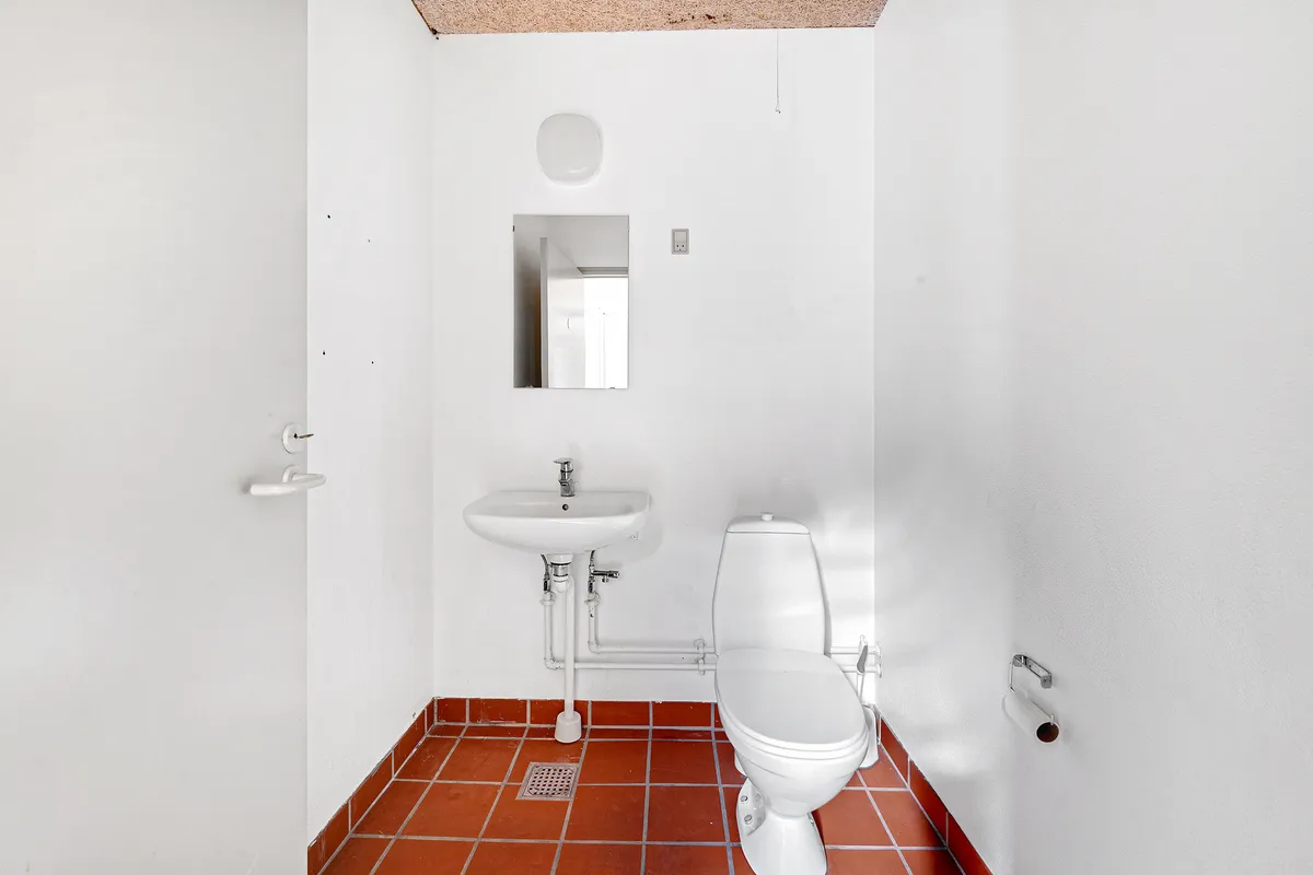 En lille toiletrum med hvid væg og rødt gulv, toilet med papirhylde og spidset håndklædeholder, samt spejl og vask med