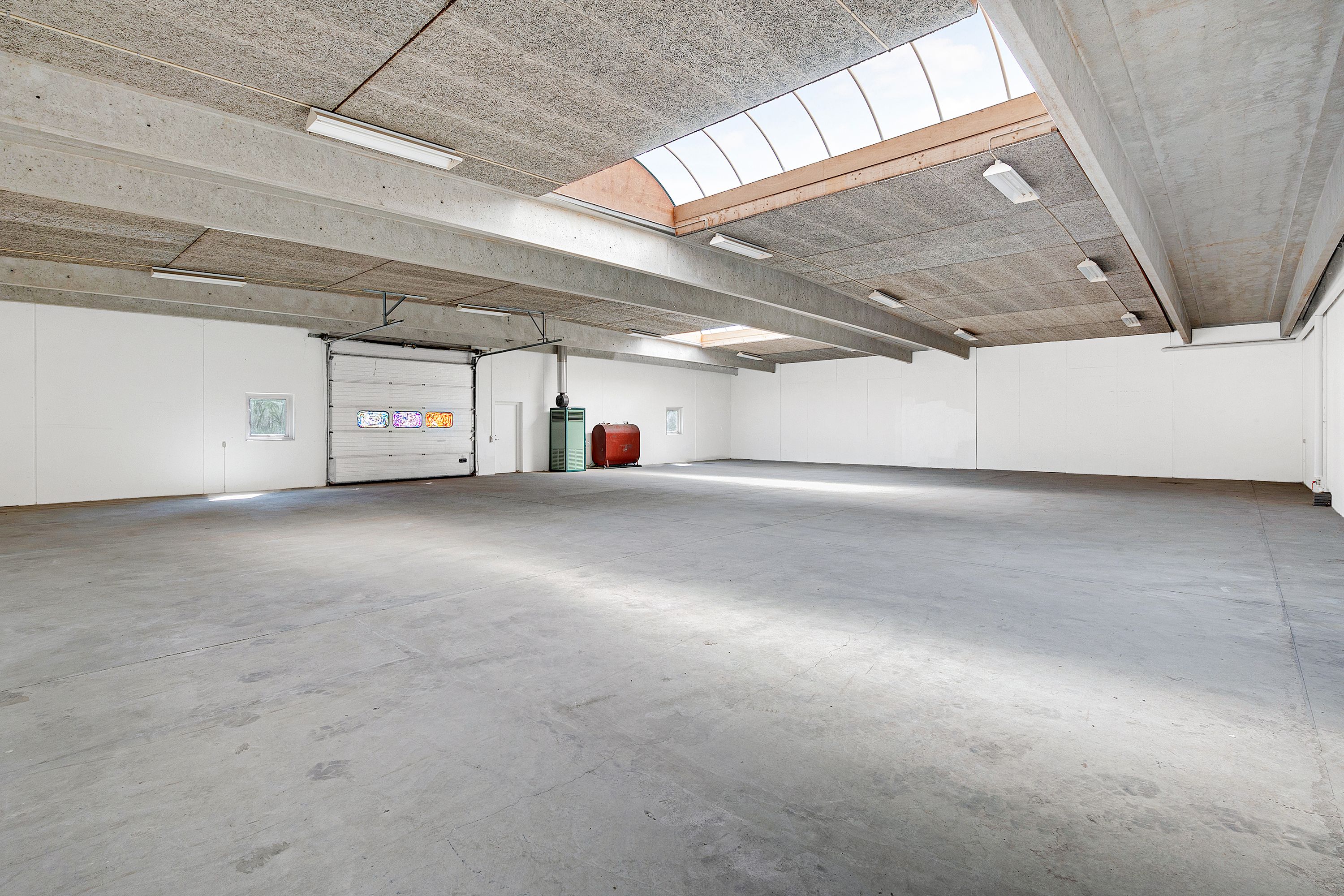 Et stort, tomt værksted med højloft, hvor der er lys, loftsvinduer, hvide vægge og en stor garagedør, der er åben.