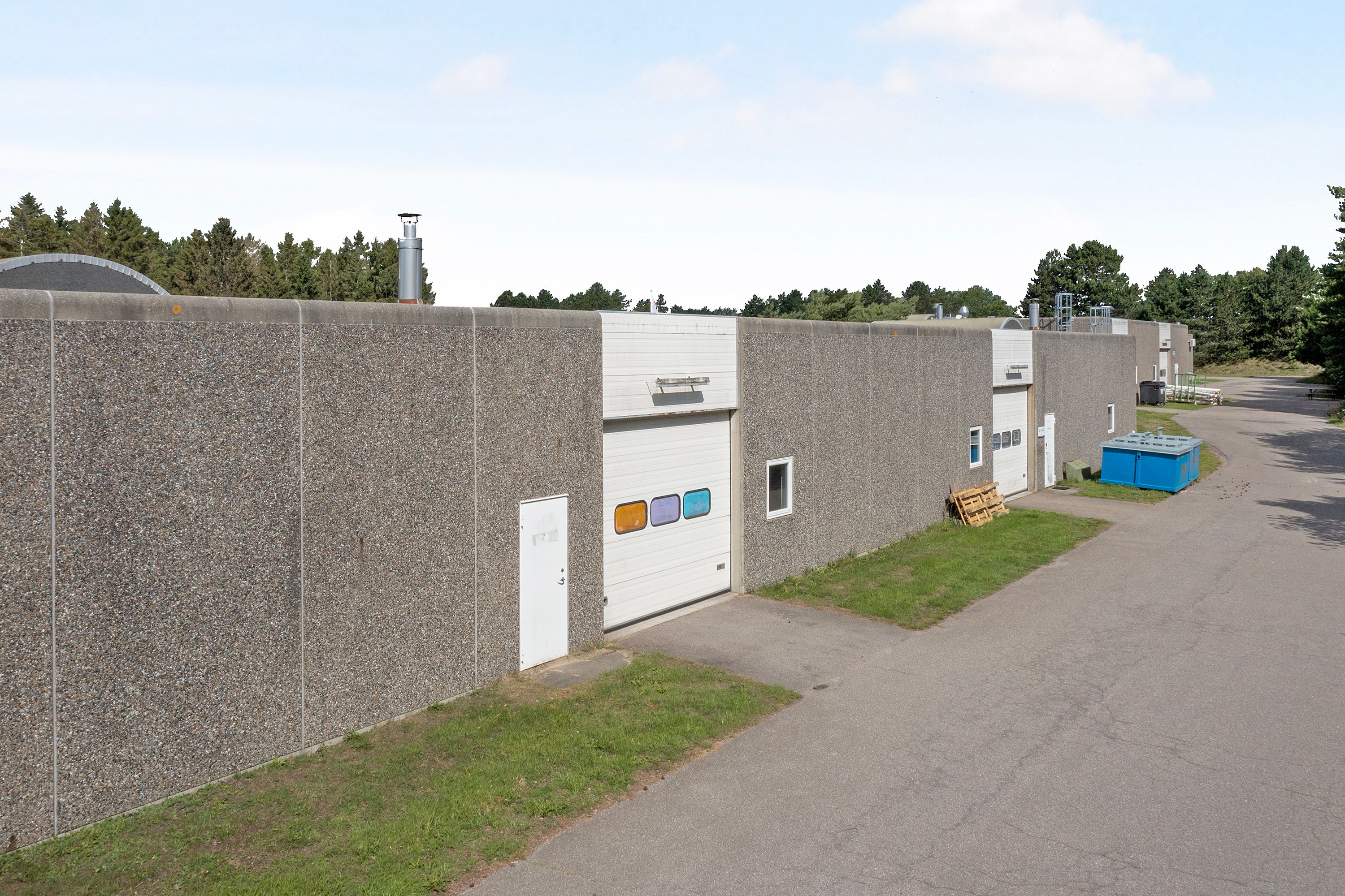 En industriel bygning med mange garagedøre og lyseruder placeret ved siden af en asfaltvej og græsarealer