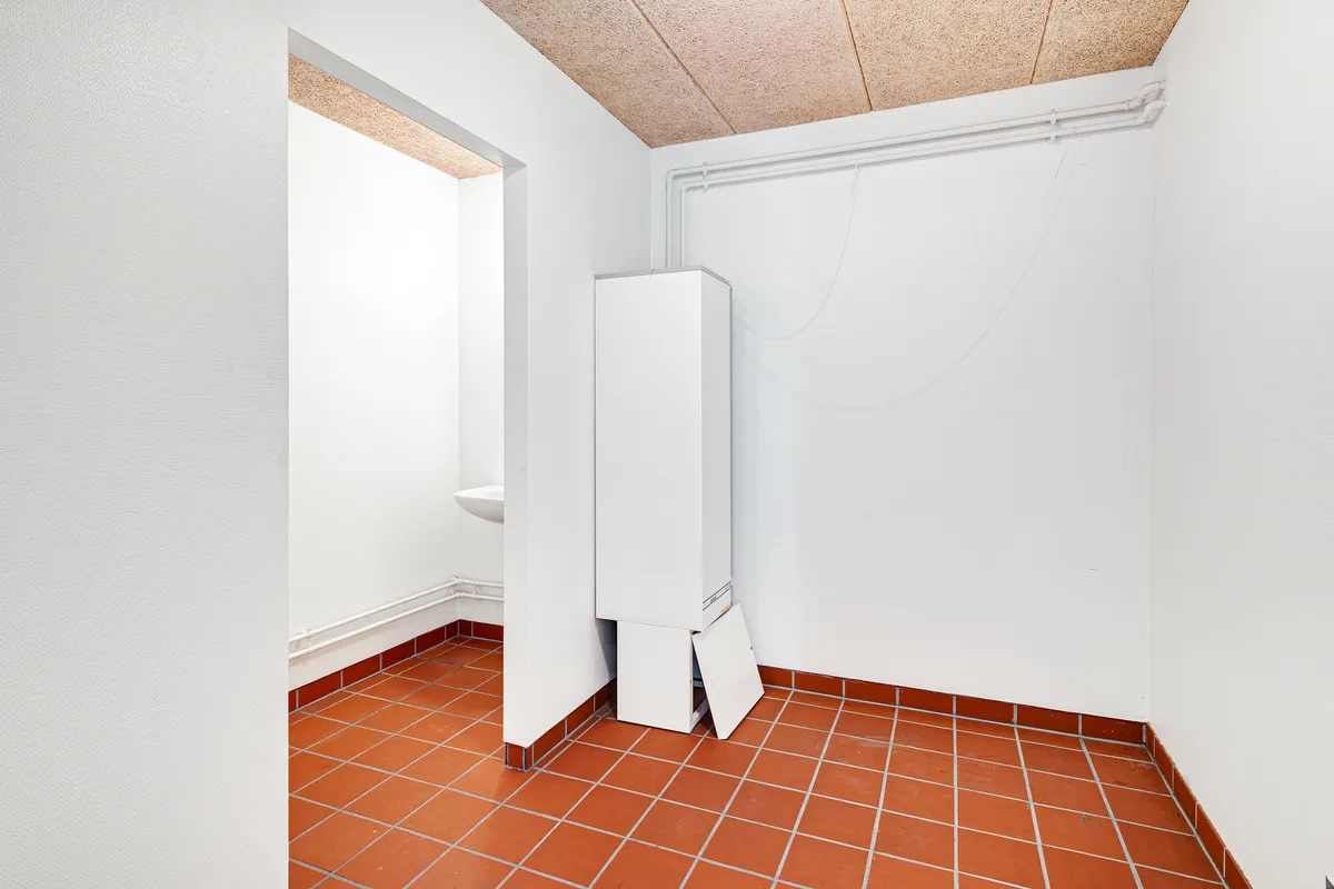 Et hjørne med hvidt vægpanel og rødgule fliser på gulvet, samt et hvidt skab med åben dør og et toilet i hjørnet