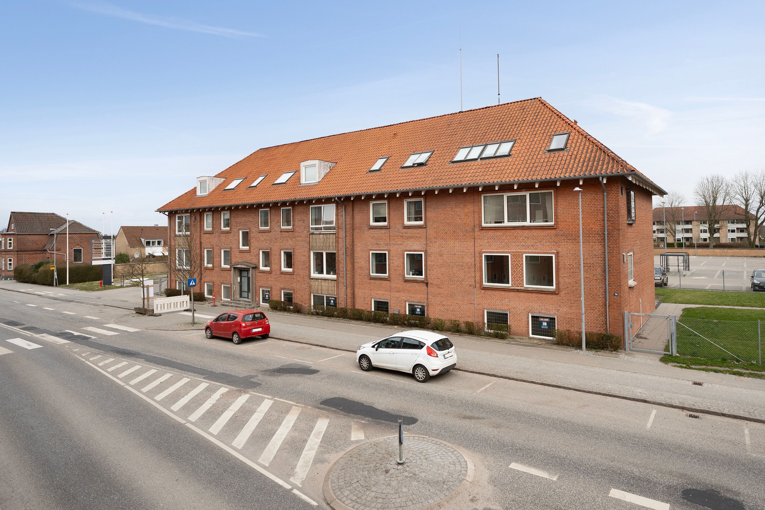 Et lejlighedshus med røde mursten og vinduer med hvide indramninger, og to biler parkeret foran