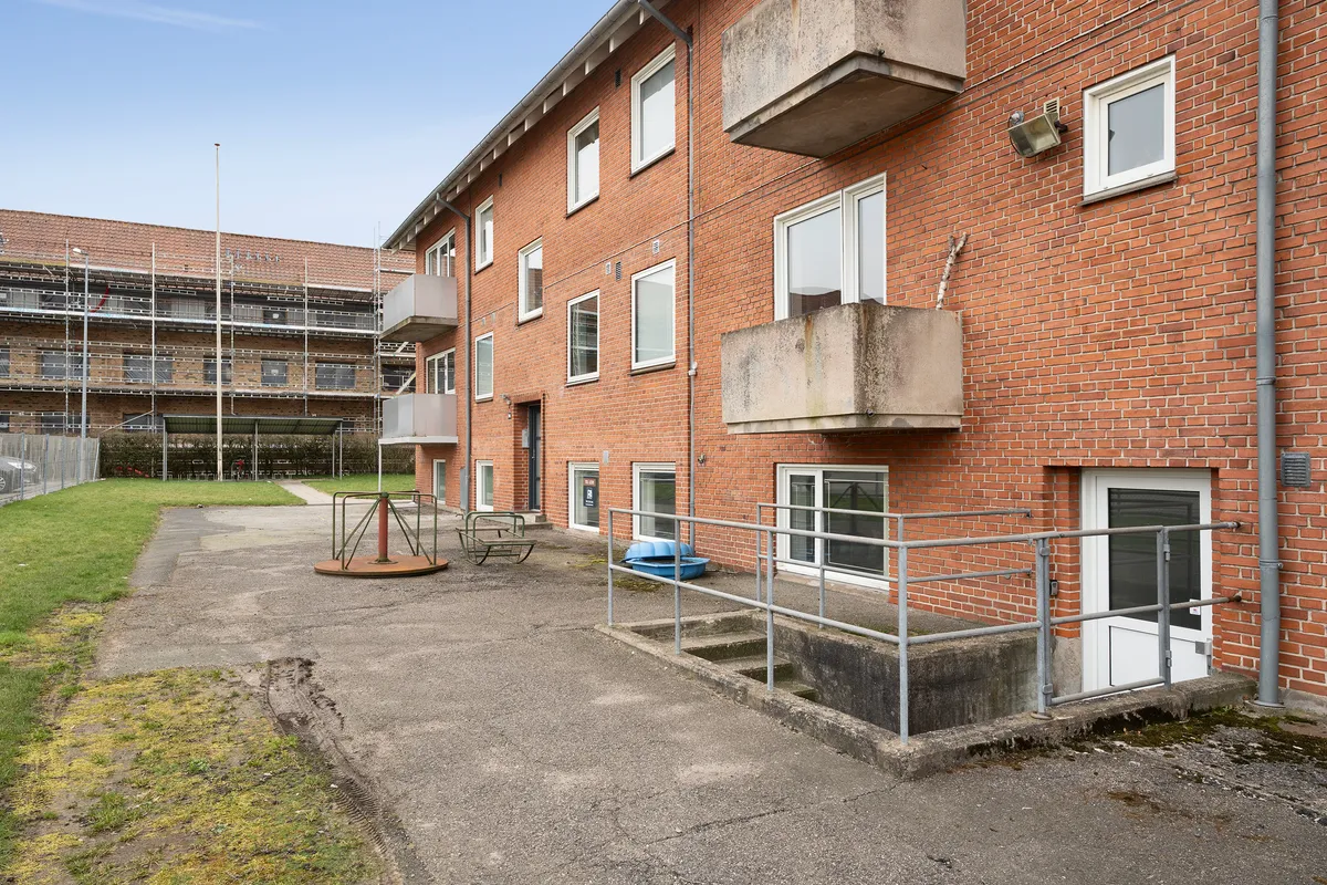 Lejlighedsbygning med rød mursten, mange vinduer, terrasser, gange og et fortov med græs.