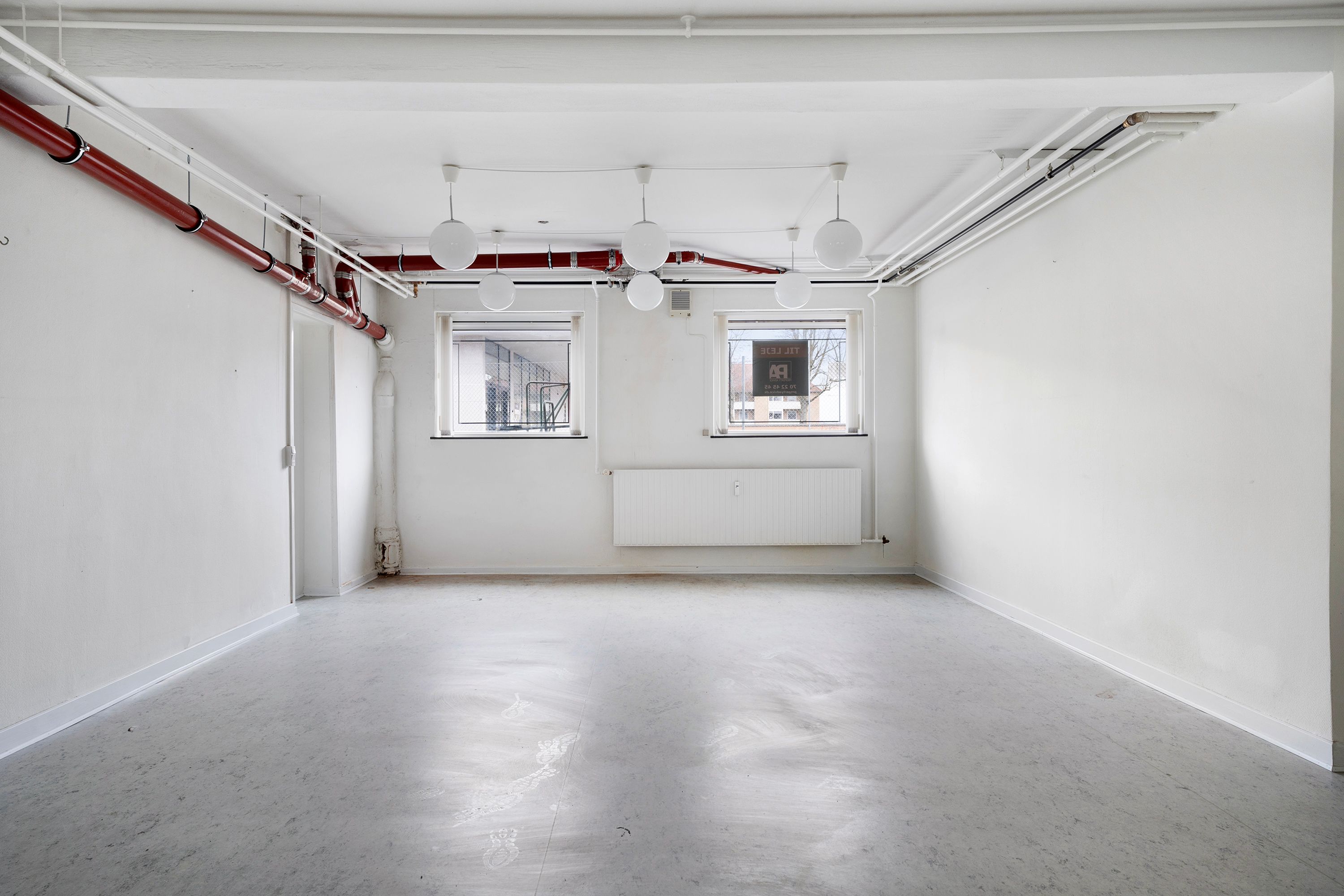 Hvid kælderlejemål med to vinduer, lysekroner, radiator, rør og gulvbelægning til renovering