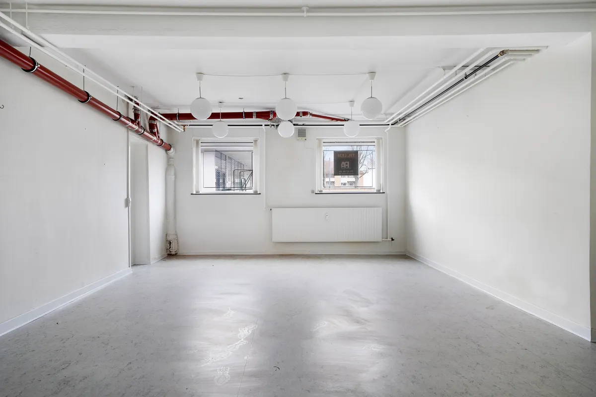 Hvid kælderlejemål med to vinduer, lysekroner, radiator, rør og gulvbelægning til renovering