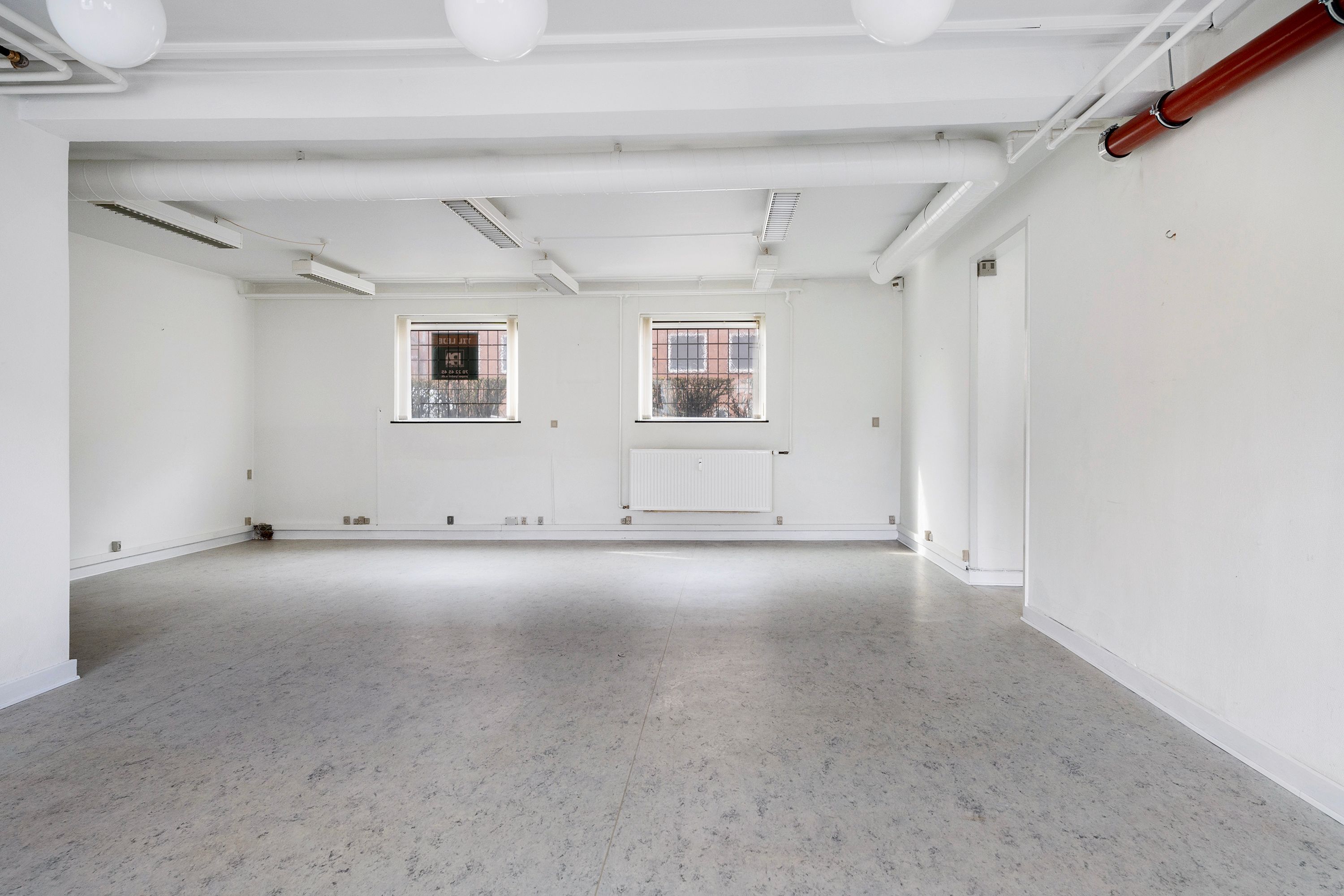 Store kælderlejemål med store vinduer og hvide vægge, lysekroner og ventilationsrør på loftet og gulvet.