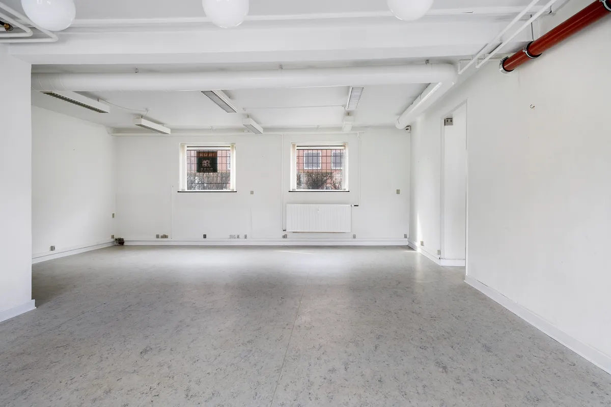 Store kælderlejemål med store vinduer og hvide vægge, lysekroner og ventilationsrør på loftet og gulvet.